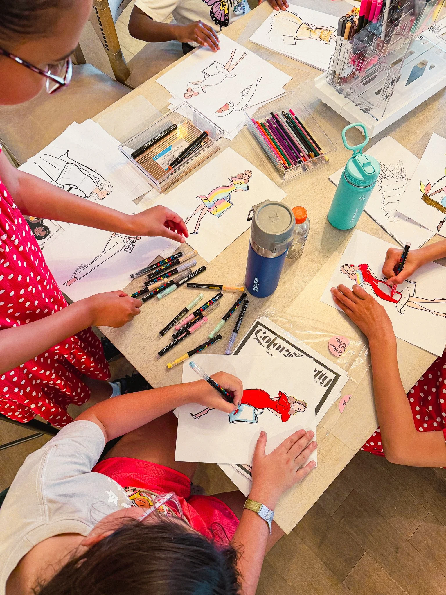 Children sitting around a table drawing and coloring fashion sketches of women with markers and colored pencils.