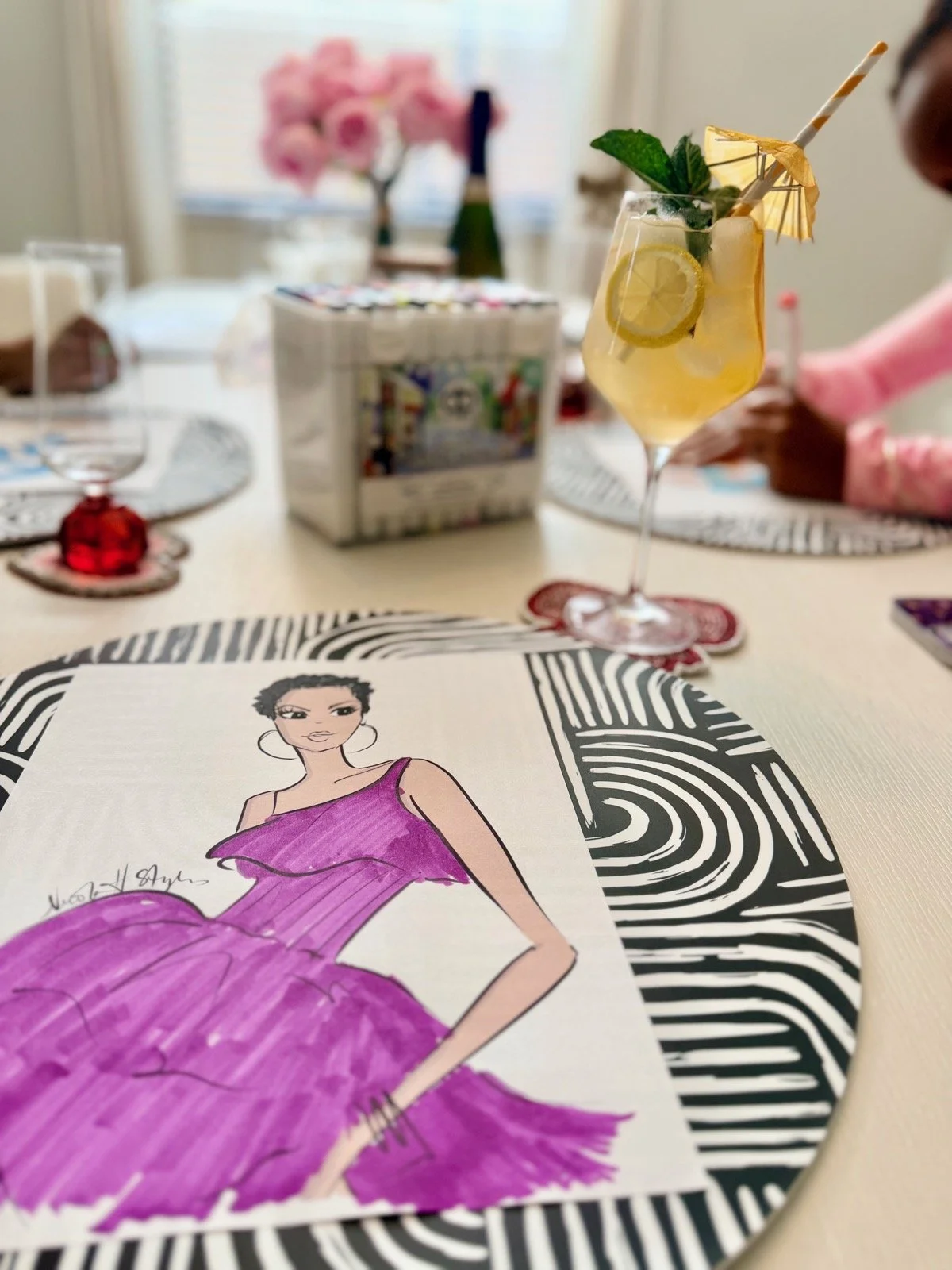 A table setting with a fashion illustration of a woman in a purple dress, a glass of lemonade with lemon slices and mint, and a blurred background including a pink flower arrangement and a box of tissues.