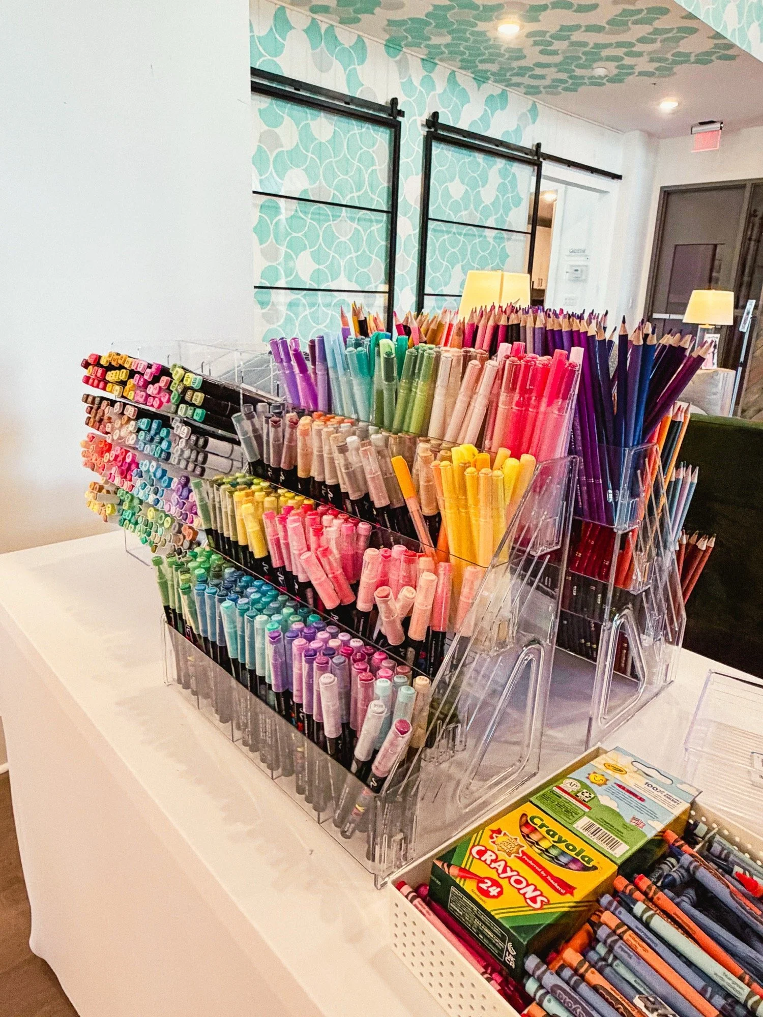 Display of colorful markers, pens, and crayons organized in clear acrylic containers on a white table, with boxes of Crayola crayons and other art supplies nearby, in a well-lit room with patterned light blue wallpaper.