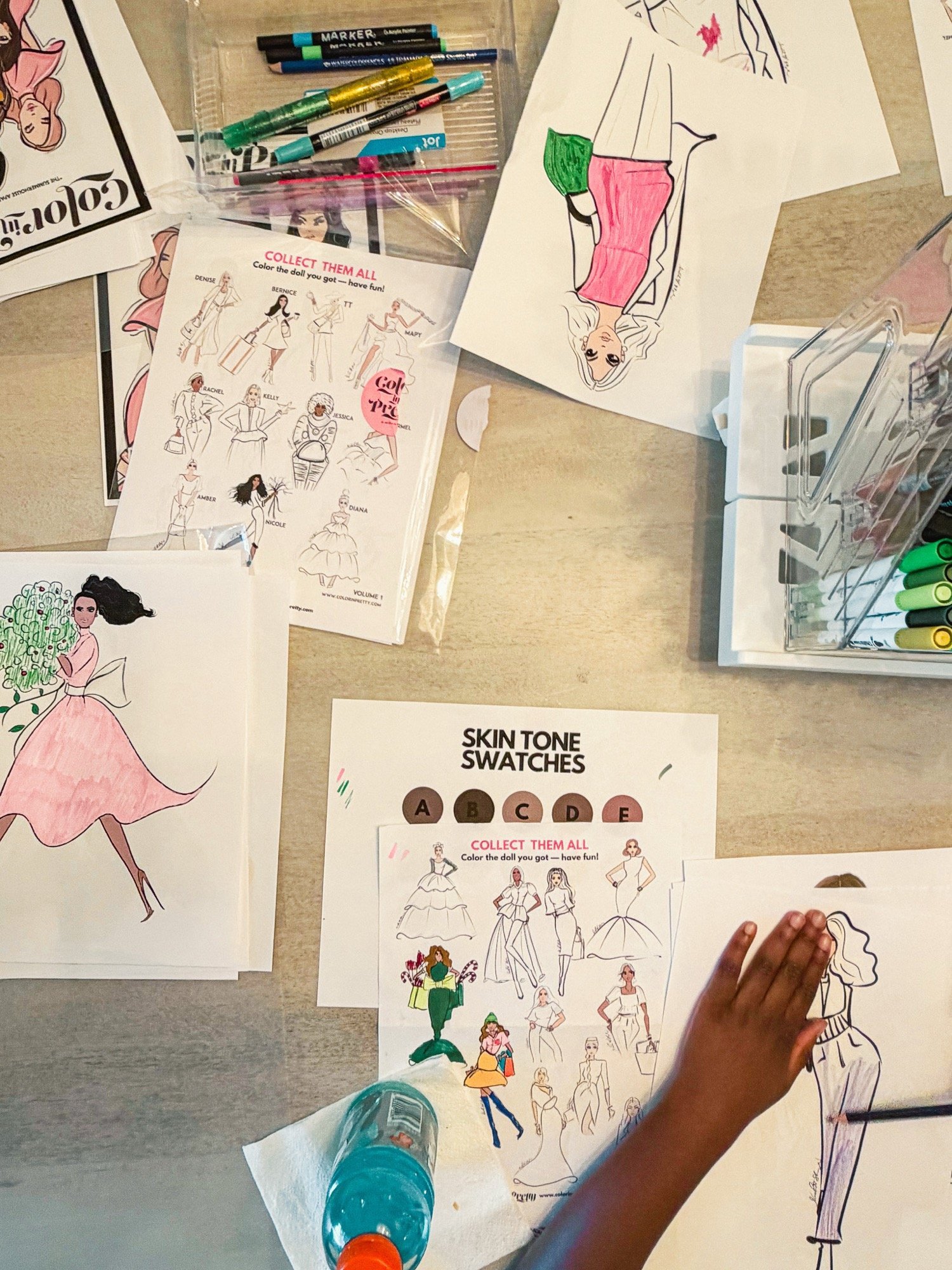 A workspace with various fashion illustration drawings and coloring sheets, including a woman in a pink dress holding a bouquet, and another with a green skirt and yellow top. There are markers and pens scattered on the table and a water bottle, with