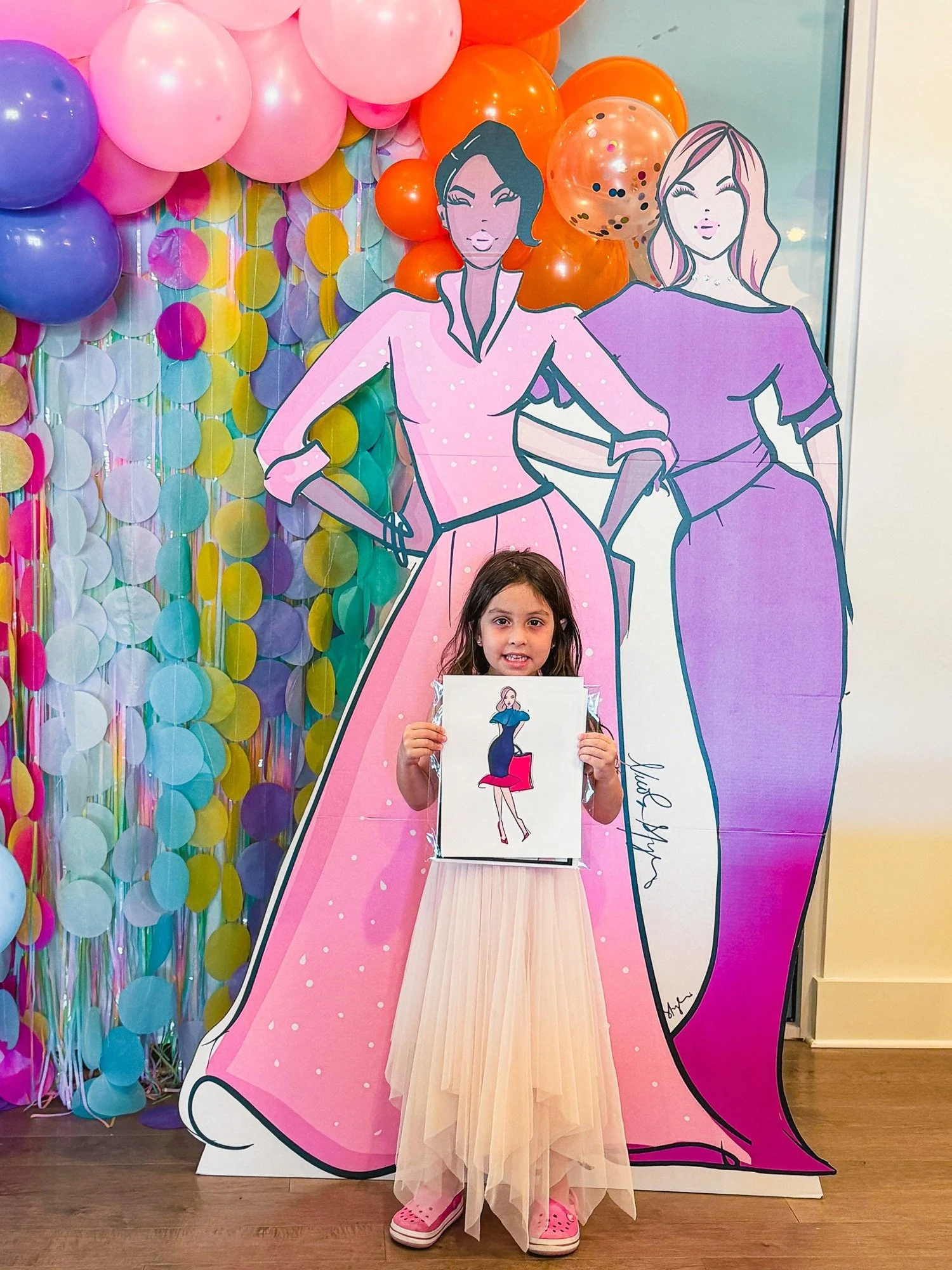 A young girl dressed in a pink shoe, beige tulle dress with a plastic bag in hand standing in front of a large pink cardboard cutout of a woman with a short bob haircut and another woman with long wavy hair, surrounded by colorful balloons including 