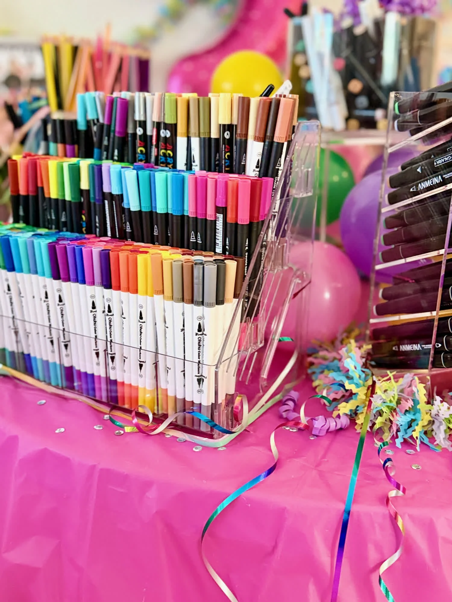 A display of colorful markers arranged in a cup, decorated with pink and purple balloons and paper streamers on a pink table.