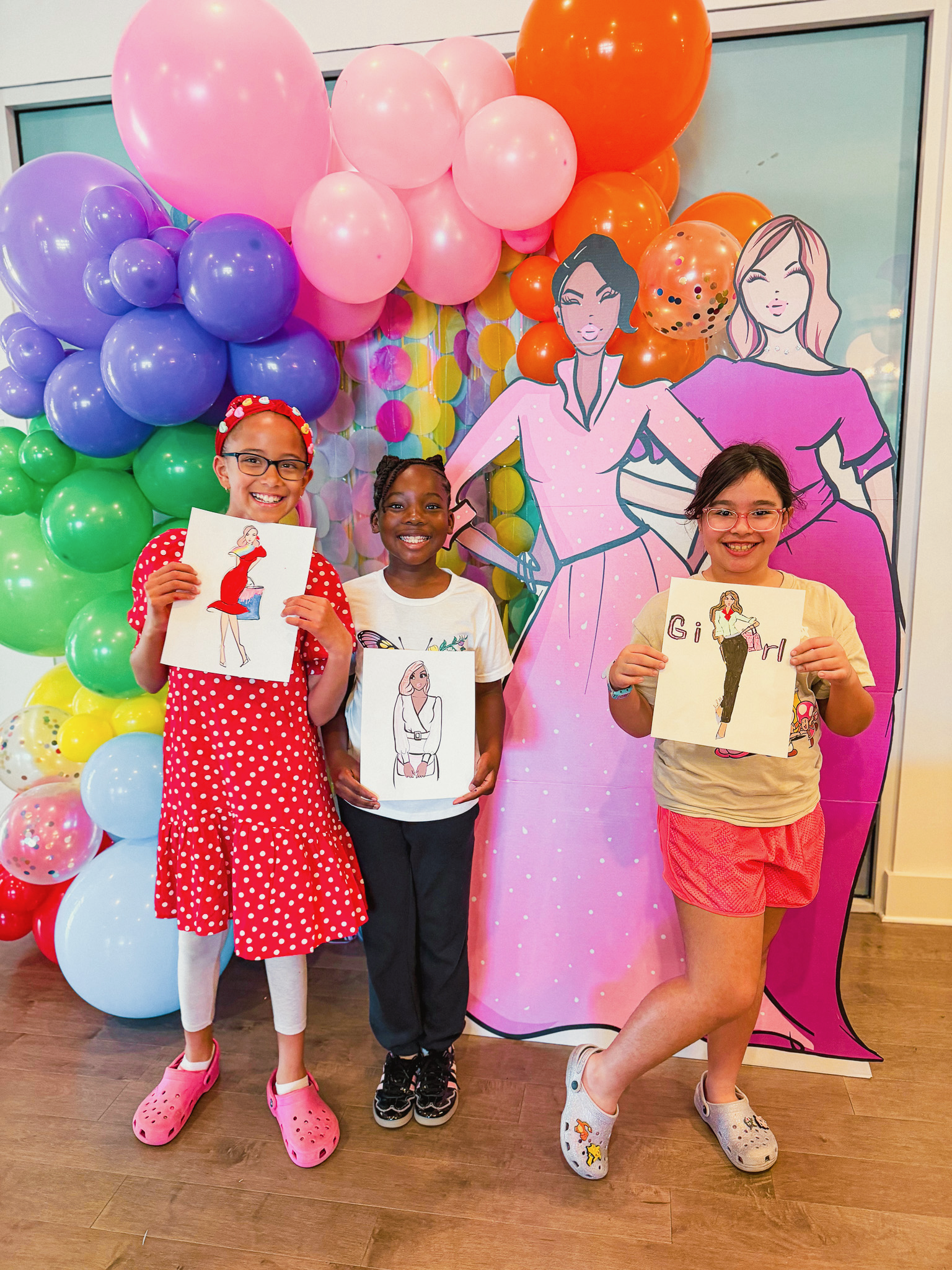 Three young girls standing in front of a colorful wall of balloons and cartoon illustrations, each holding a drawing of a woman with different styles and outfits.