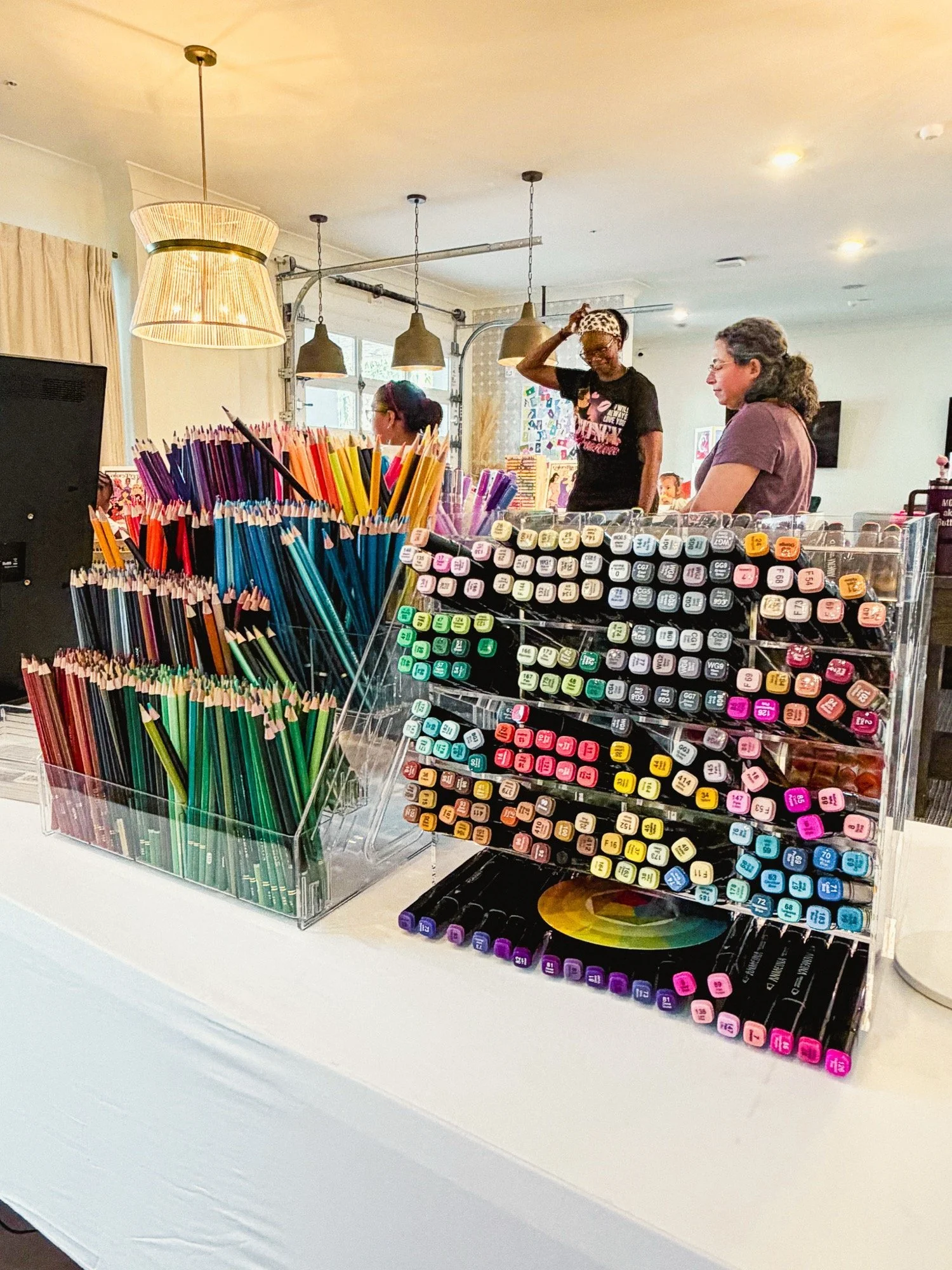 Display of colorful markers, pens, and art supplies on a table at an art store, with three women browsing in the background.