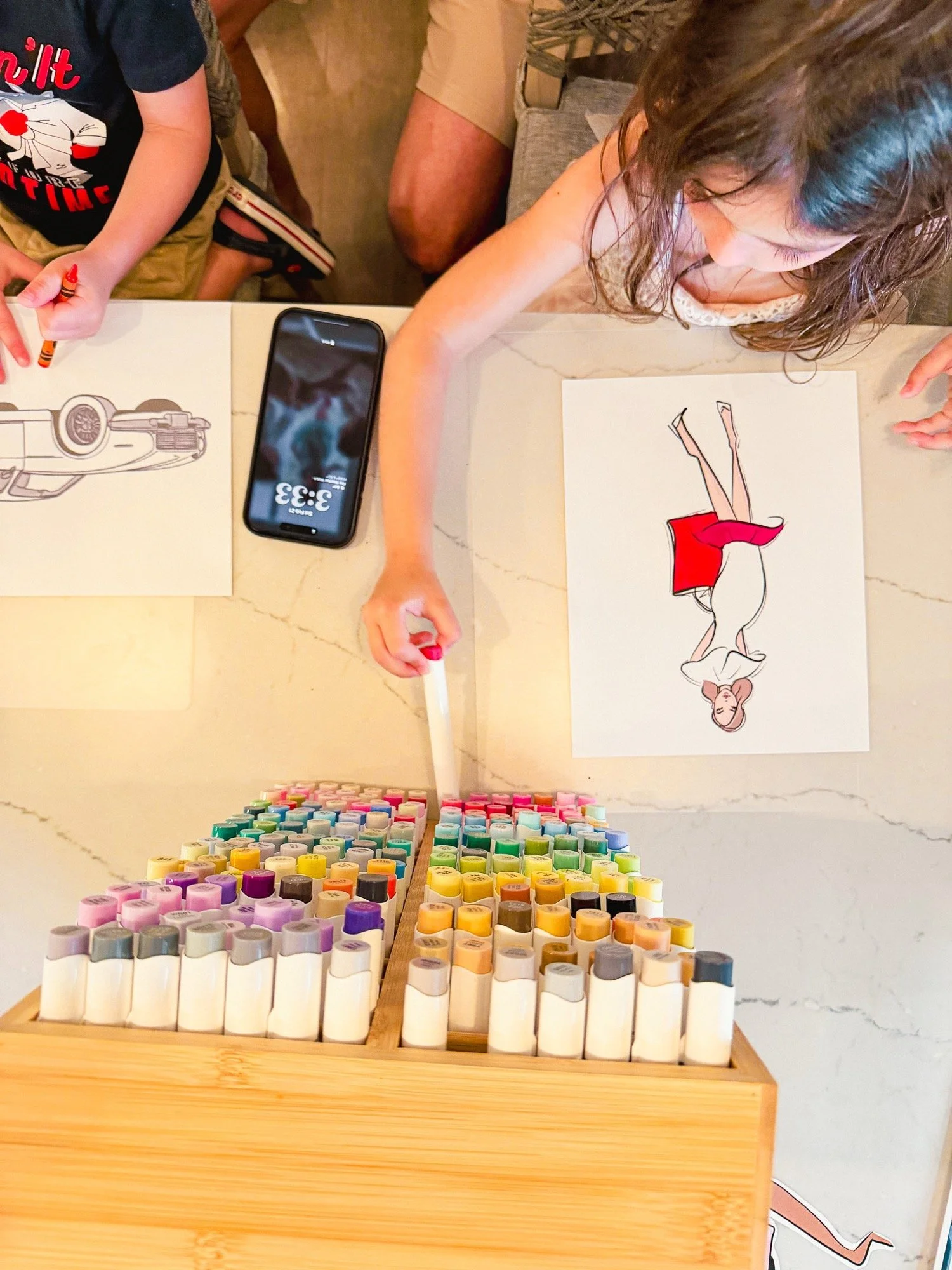 A wooden table displays rows of colorful markers, with art supplies and a drawing of a woman in a red dress on the table. A smartphone is placed on the table, reflecting the scene.