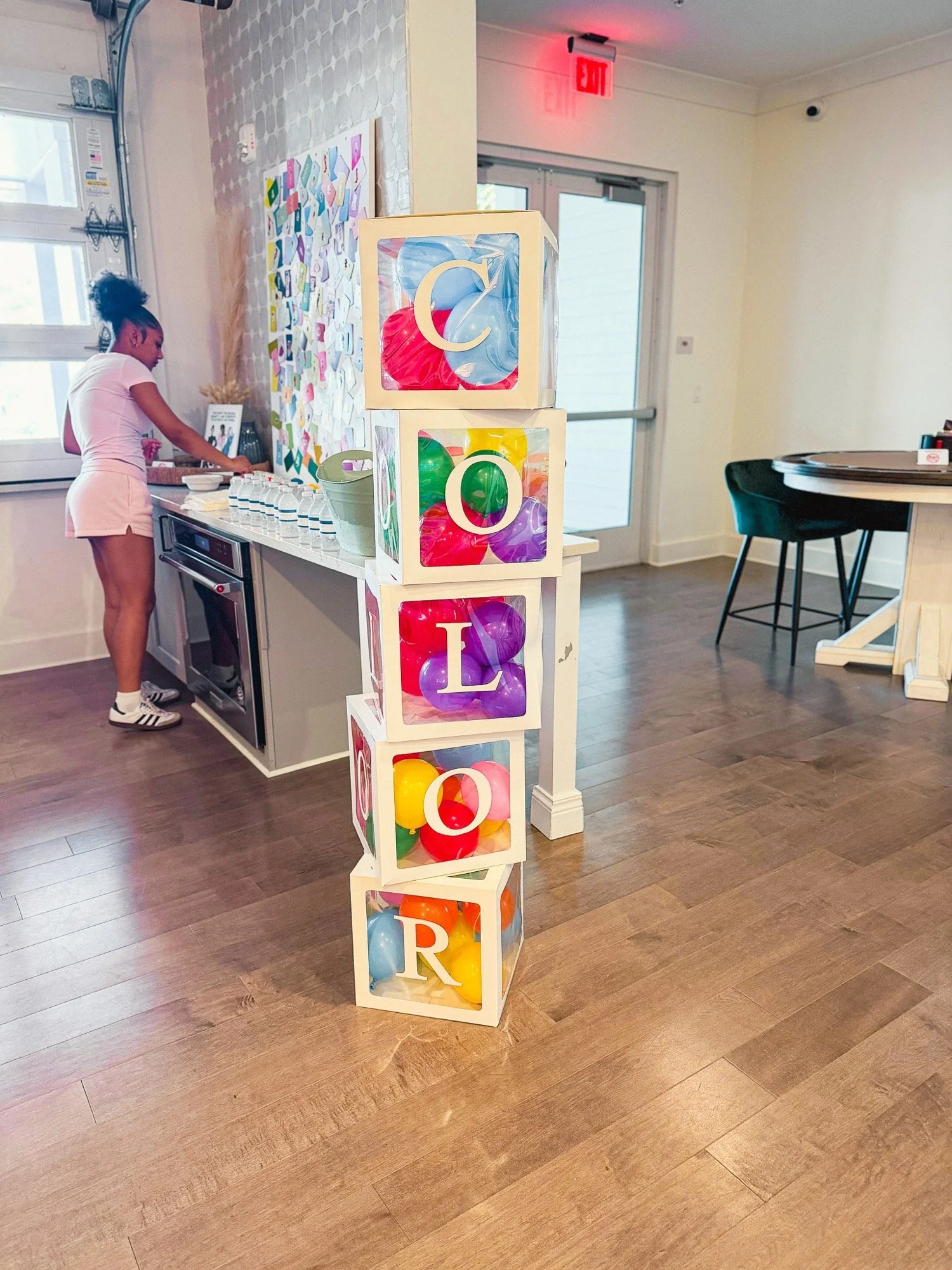 Stacked cubes with colorful balls and large alphabet letters spelling 'COLOR'. A woman is in the background preparing items in a kitchen or cafe setting with wooden floors and a white wall.