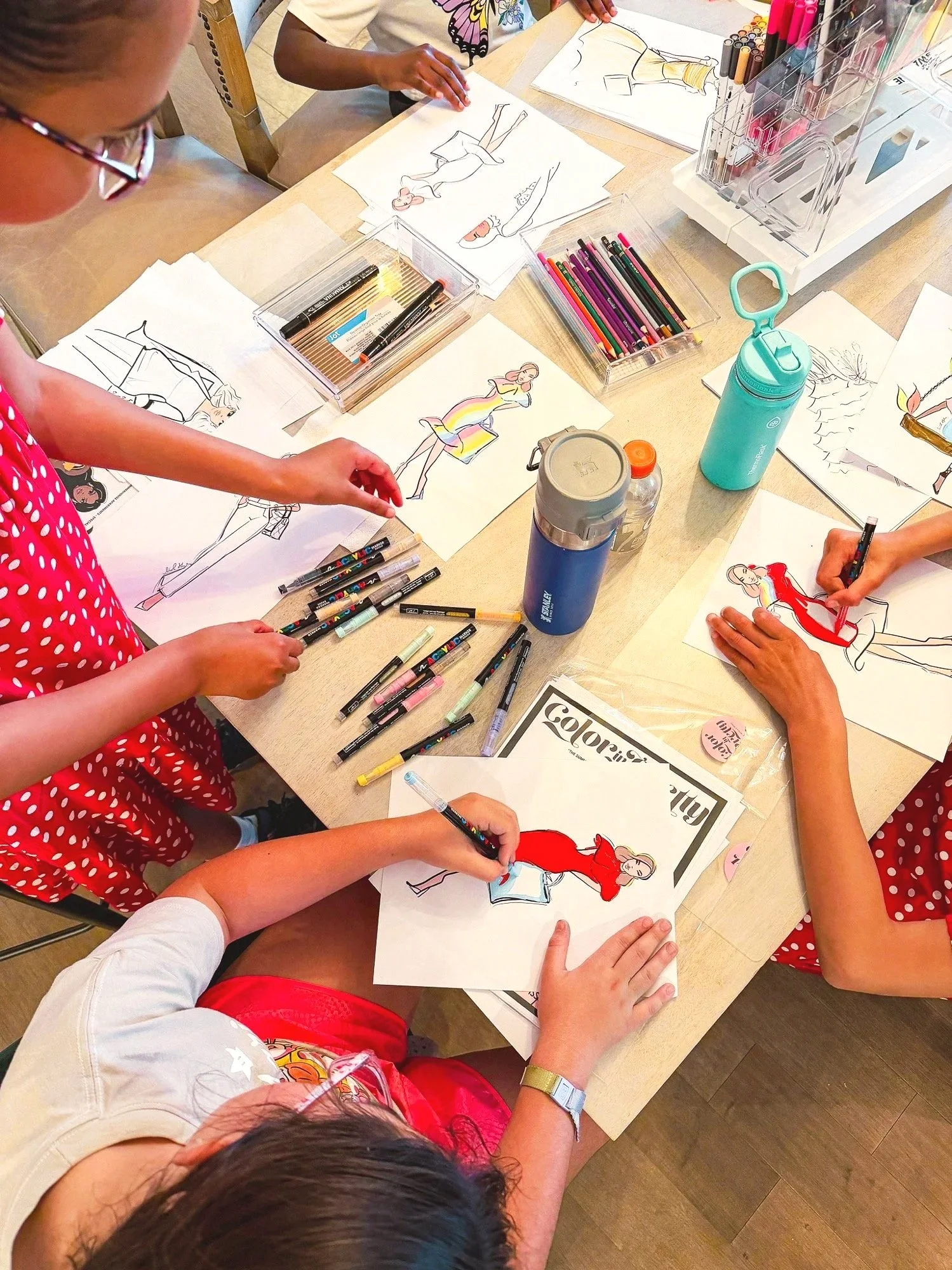 Children and adults gathered around a table coloring and drawing fashion sketches using markers and colored pencils.