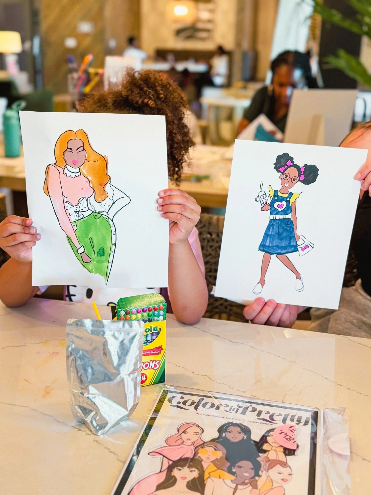 Children holding up illustrated fashion magazine pages at table in a cafe or restaurant, with coloring supplies and a magazine on the table.