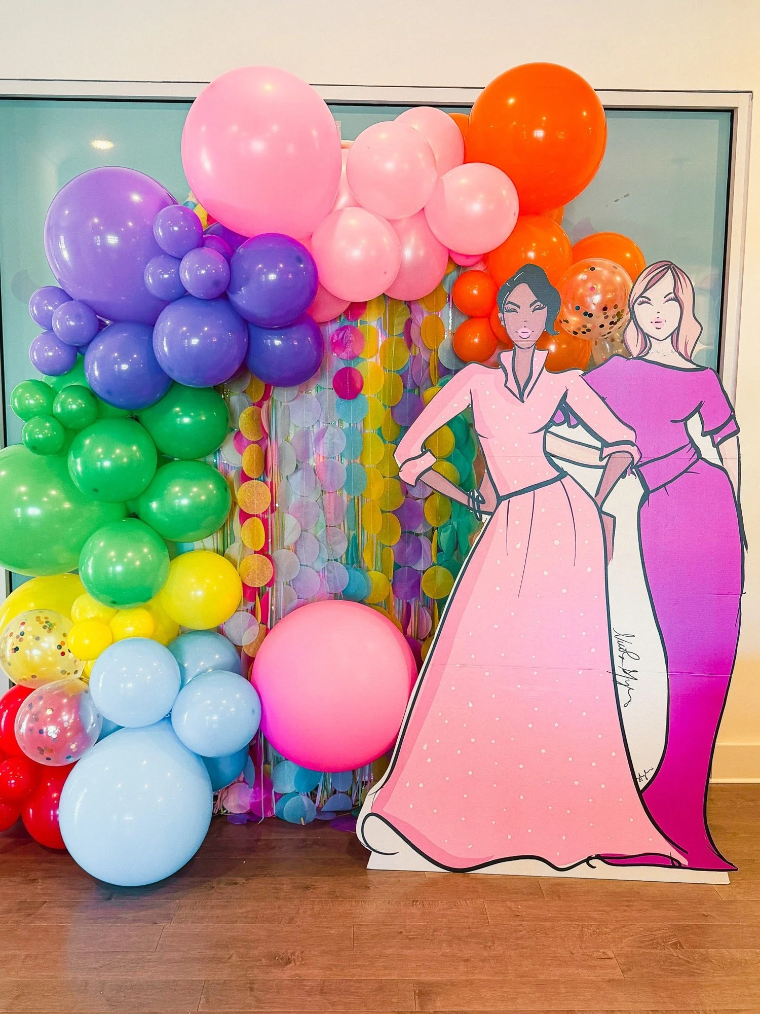 Colorful balloon display with pink, purple, green, yellow, light blue, red, and orange balloons arranged in clusters, with a large pink balloon and a cutout illustration of two women in front of the display.