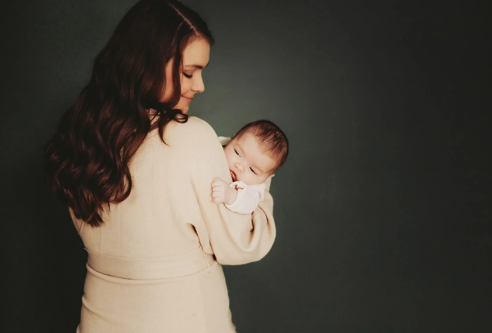 A woman with long, wavy brown hair holding a baby against a dark background.