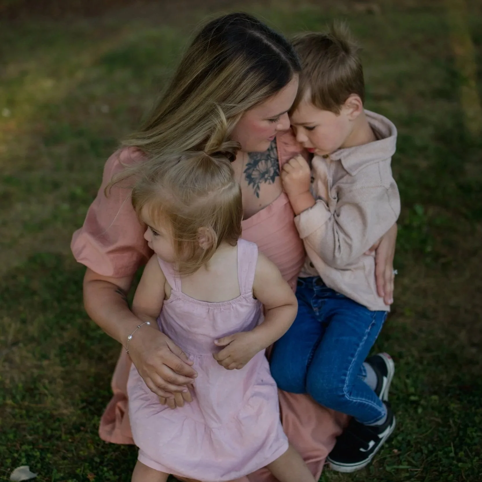 A woman with blonde hair and a tattoo on her shoulder, sitting on grass while hugging two young children, a girl in a pink dress and a boy in a light hoodie. The three are closely embracing, with their foreheads touching.