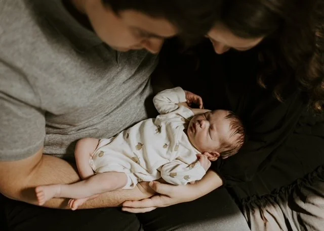 A newborn baby lying in the arms of two adults, one male and one female, who are looking at the baby and smiling.
