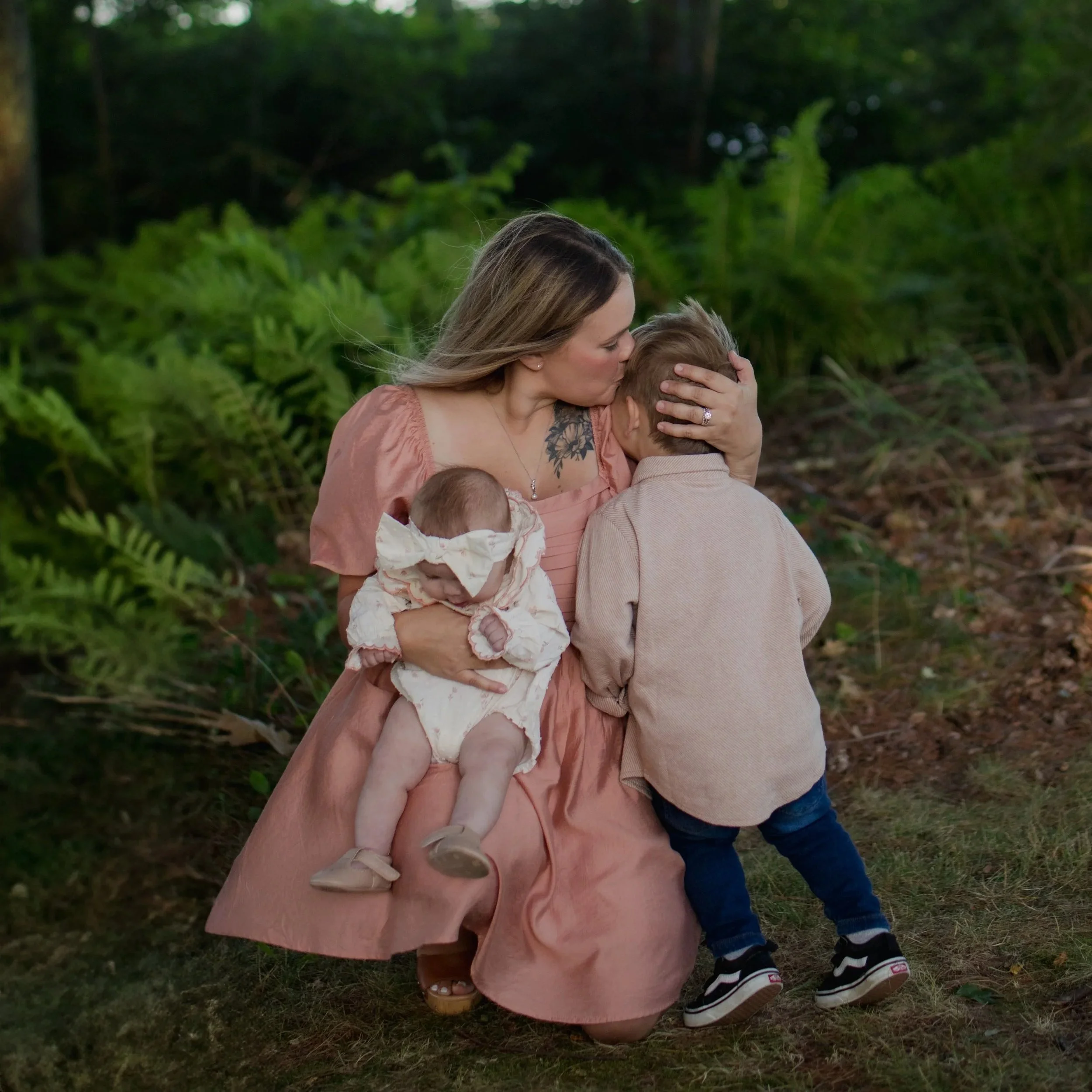 A woman with long hair holding a child in an outdoor setting with greenery, kissing the child's forehead.