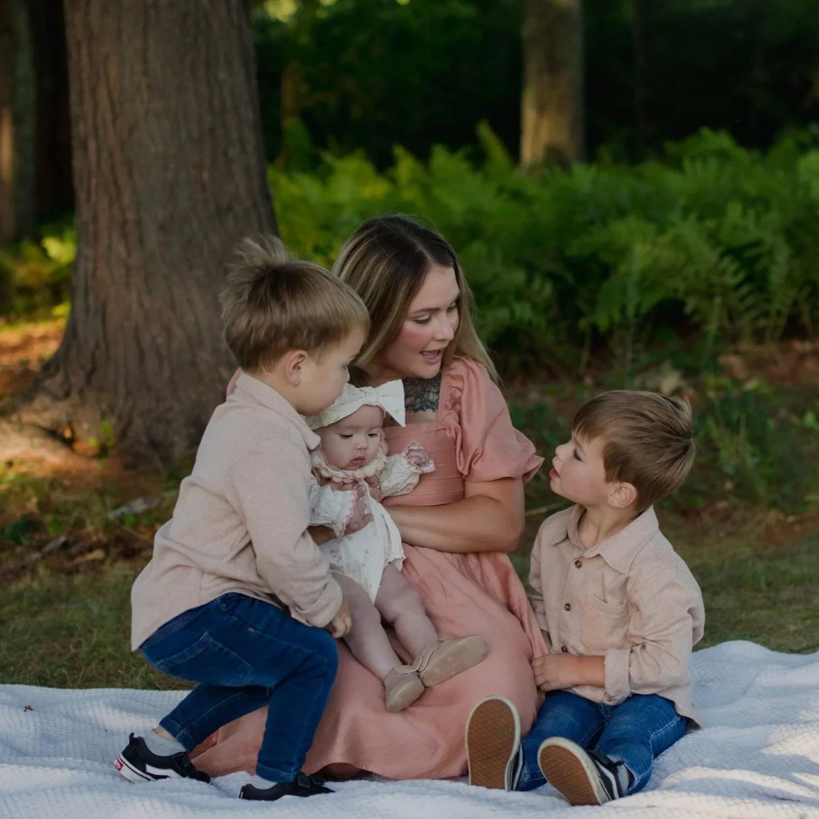A woman with long hair and four children sitting outdoors on a blanket near a large tree, engaging in conversation and interaction in a wooded area.