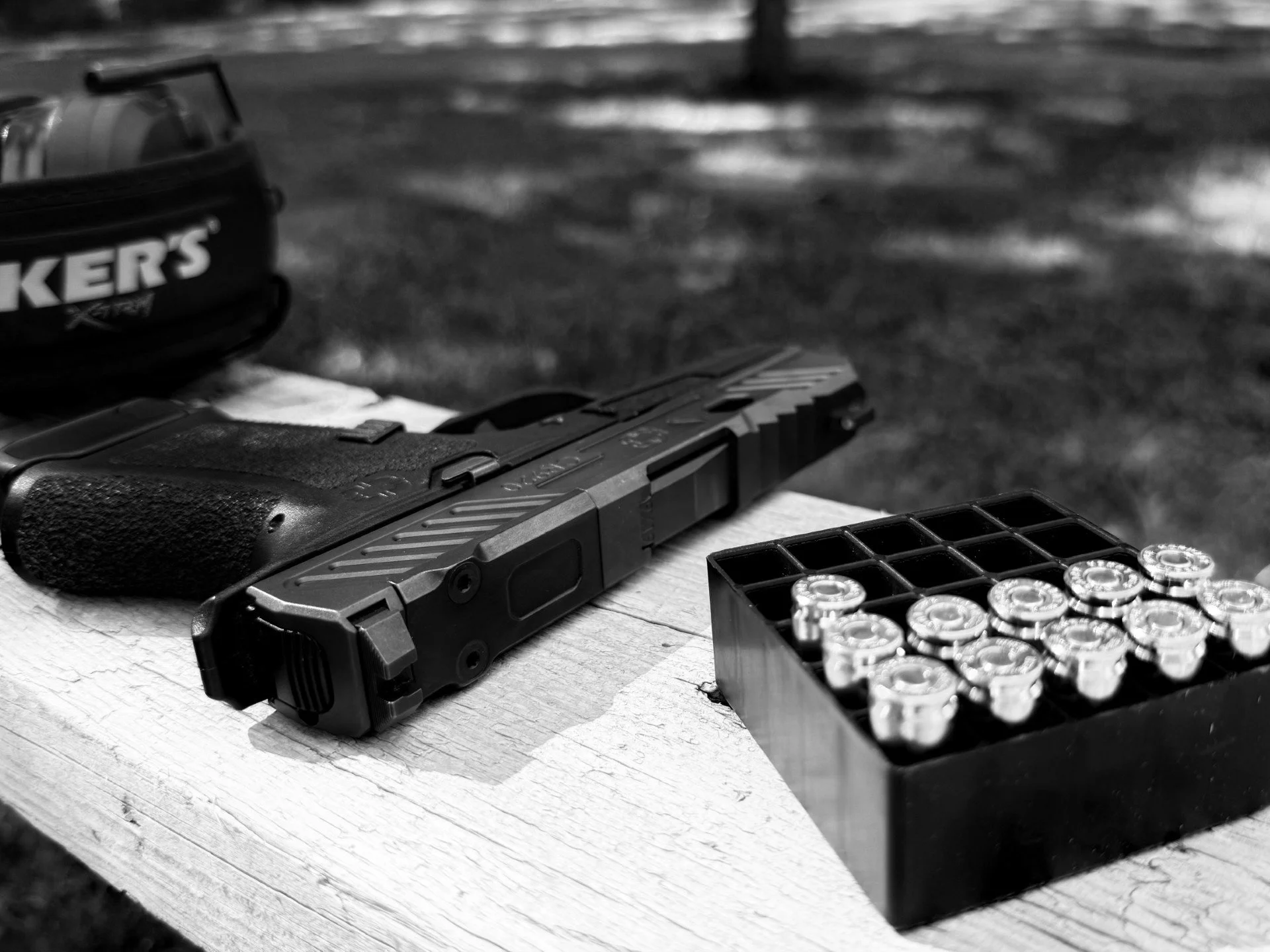A handgun, a box of ammunition with metal cartridges, and a partial view of a gun cleaning kit on a wooden surface outdoors.