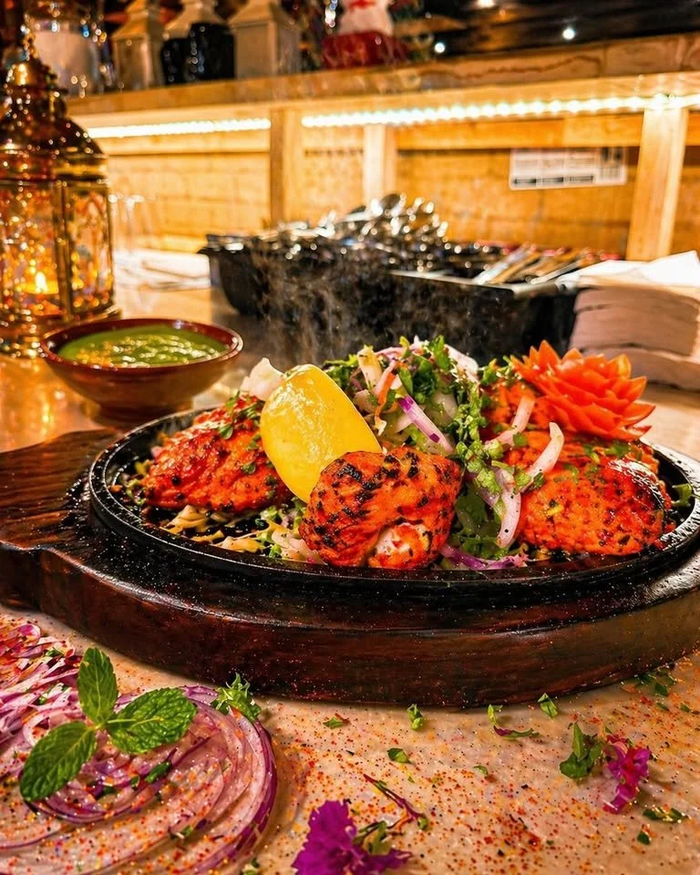 Grilled chicken topped with herbs served on a sizzling platter with lime, salad, and vegetables, in a cozy restaurant setting.