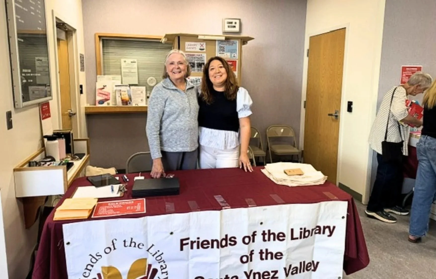 Two women standing behind a table with a maroon and white tablecloth at a library event. They are smiling and posing for the camera, with several chairs behind them. The table has flyers, a black laptop, and a stack of paper. There are other people i