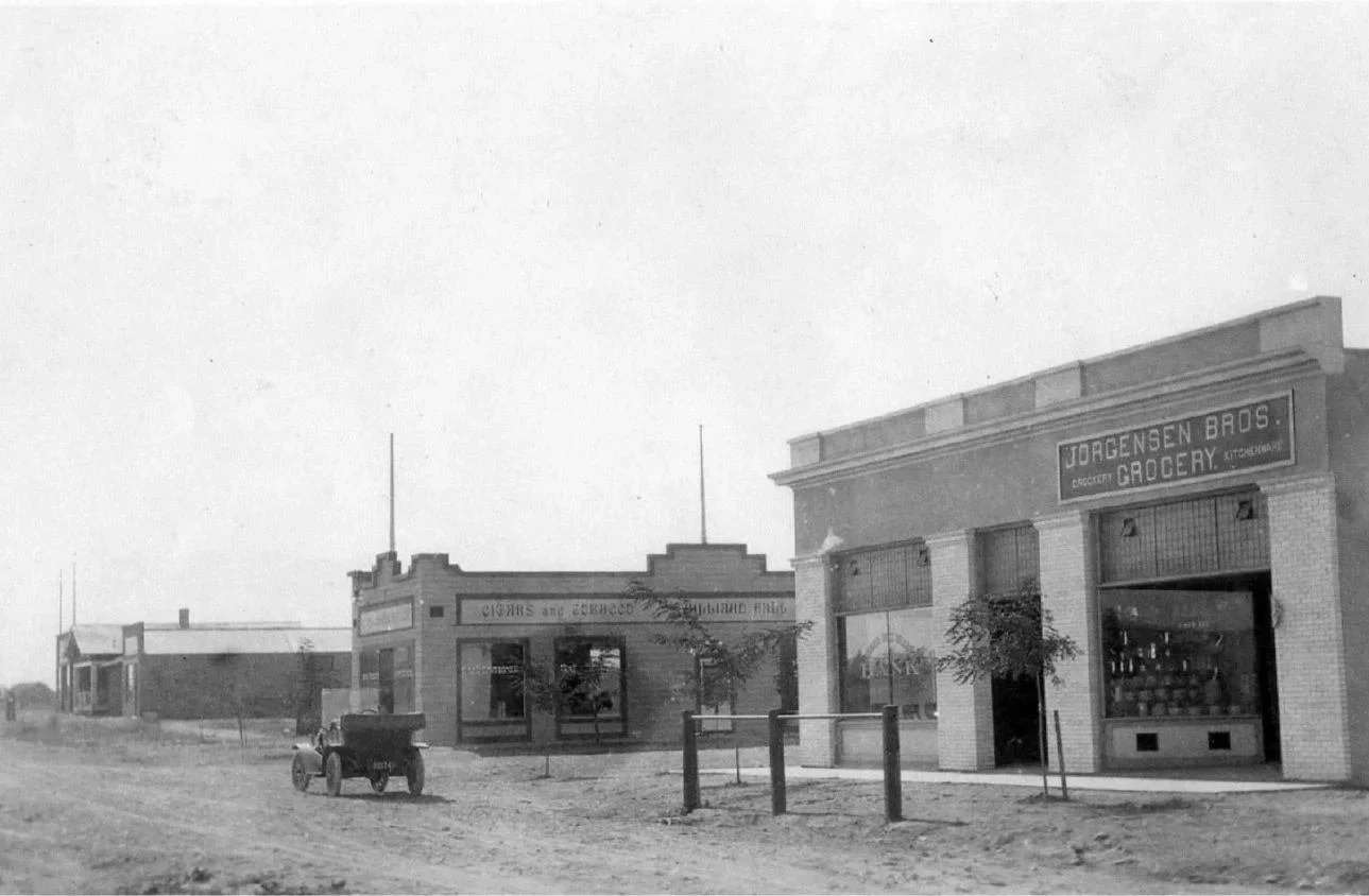 Black-and-white photo of a historical street scene in Solvang, CA with a library and small shops, including a grocery store named Jorgensen Bros. with a sign, and a nearby building with a sign reading 'Oasis and Exchange'.