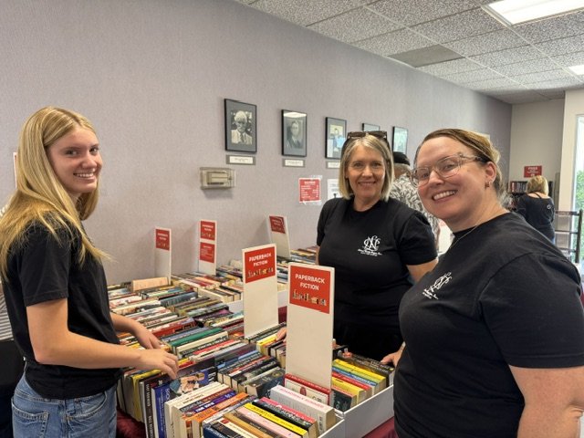Three women shopping for paperback fiction books at a bookstore or library table.