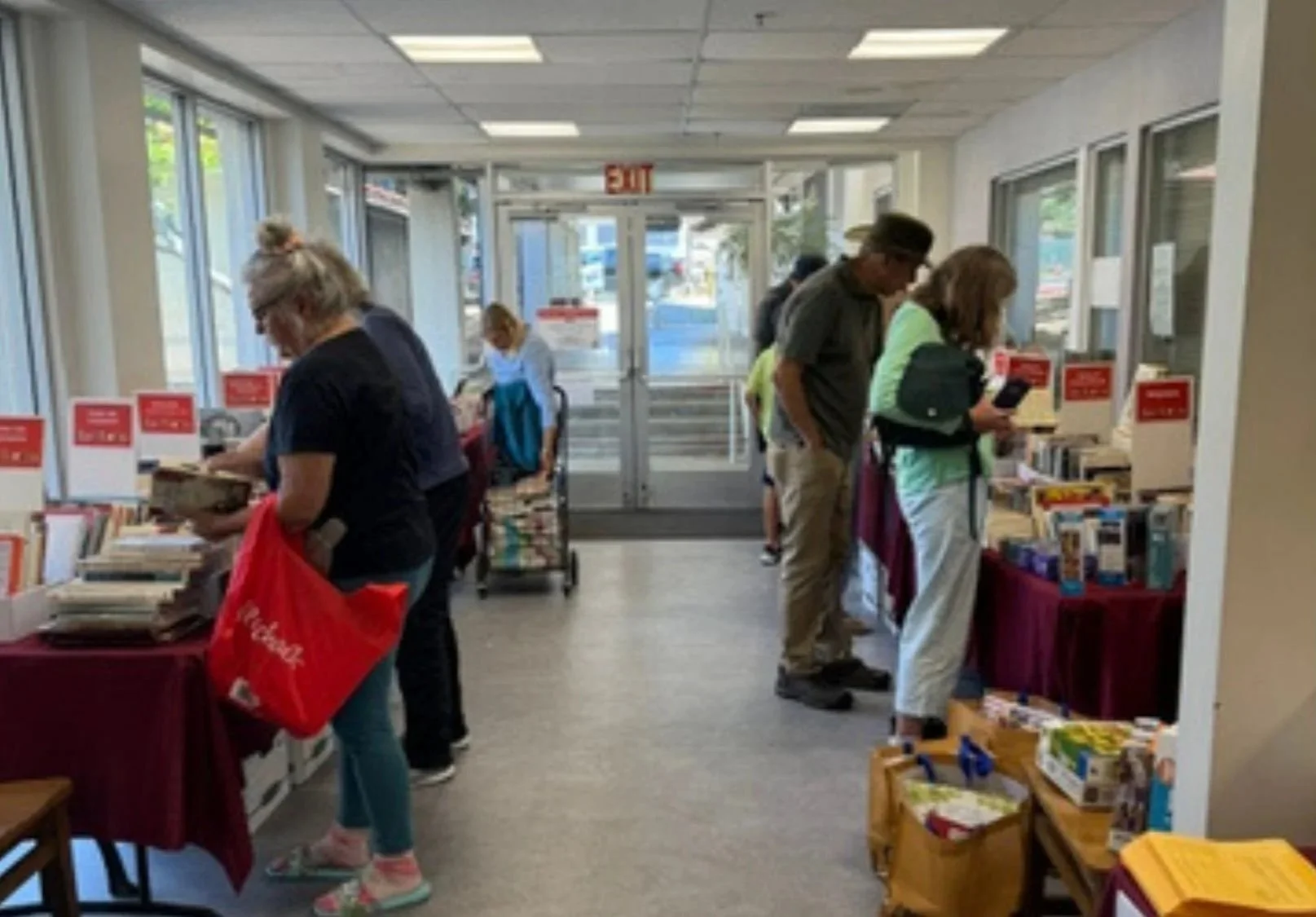 People shopping at a book sale inside a store, browsing through tables of books, with a glass entry door and windows visible in the background.