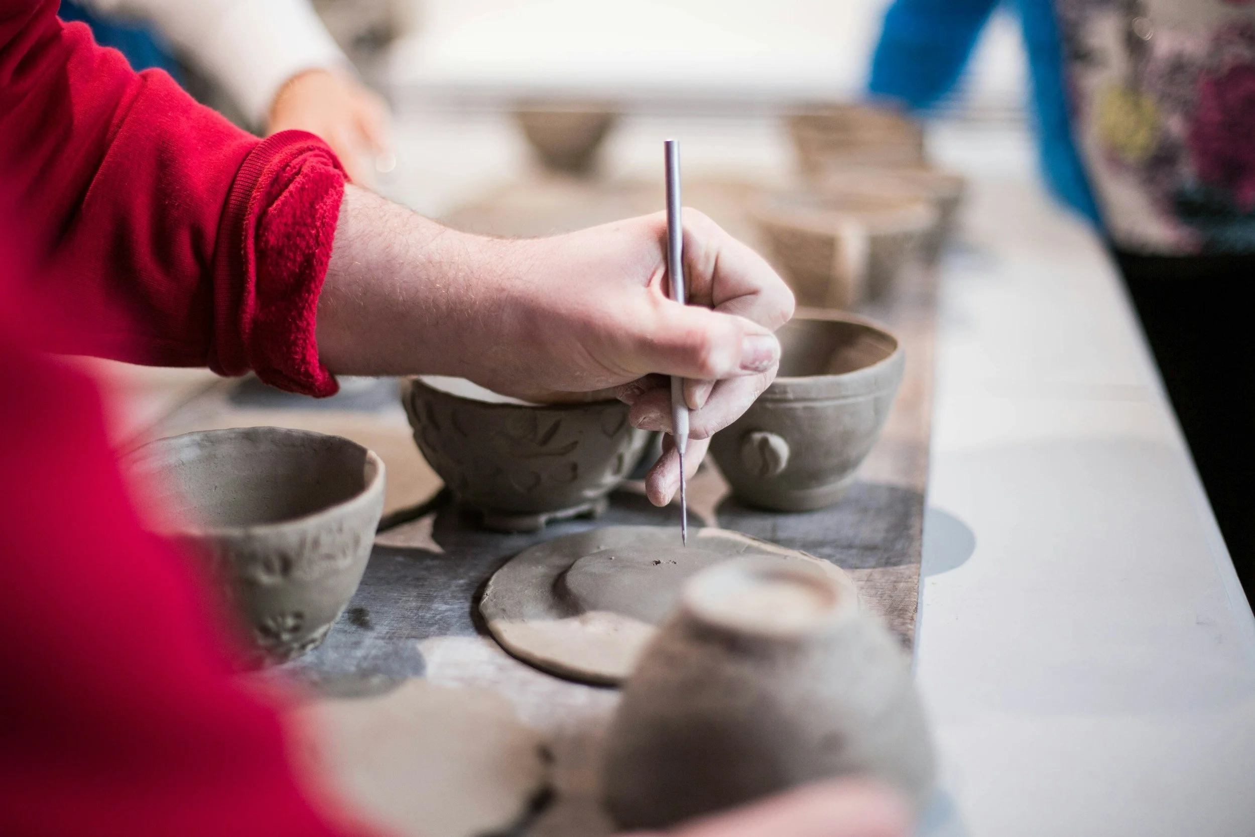 Person in a red sleeve shaping and carving clay bowls on a pottery wheel with other pottery bowls nearby.
