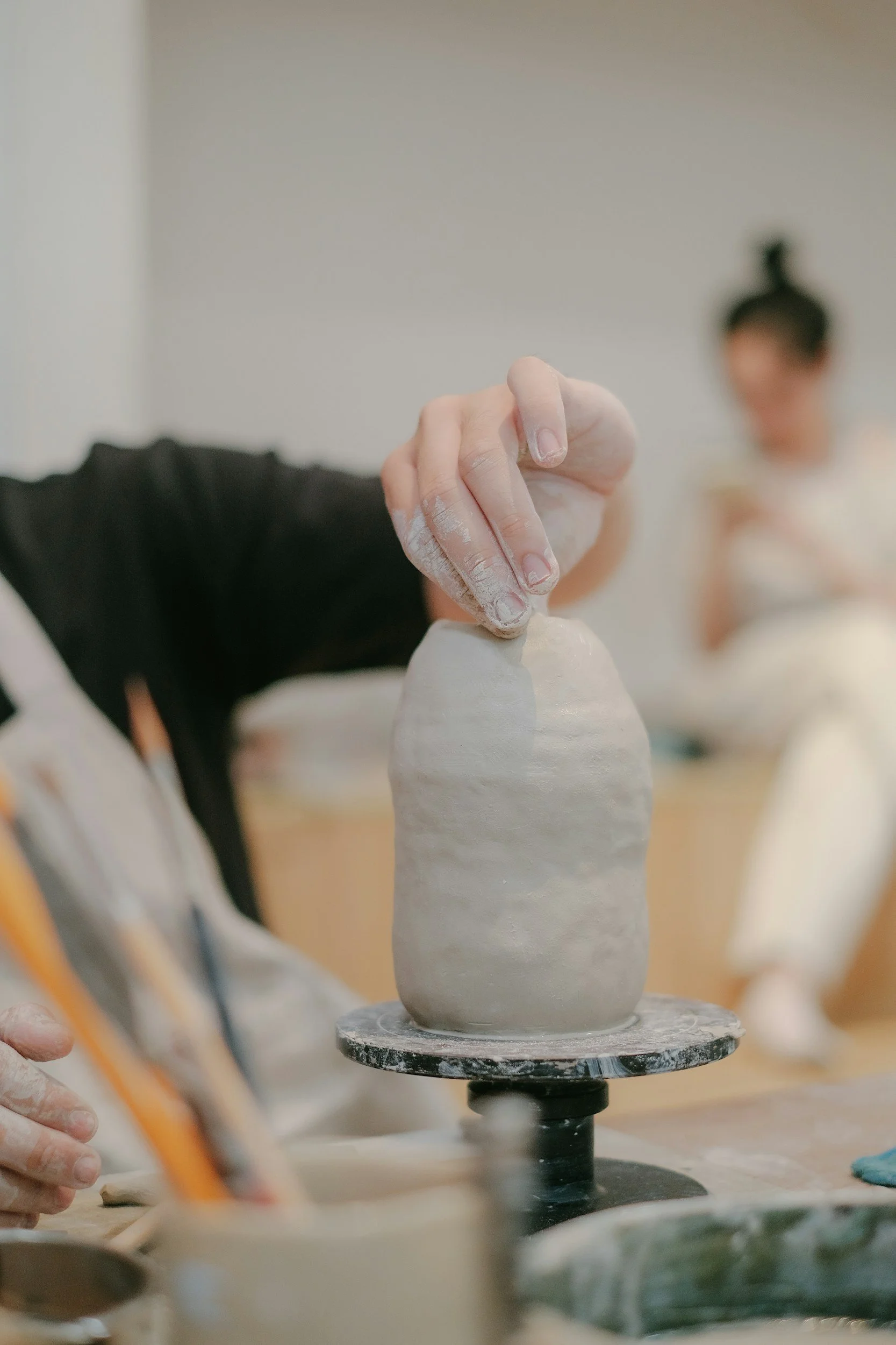 A person shaping a ceramic sculpture on a pottery wheel with their hand covered in clay, while another person watches in the background.