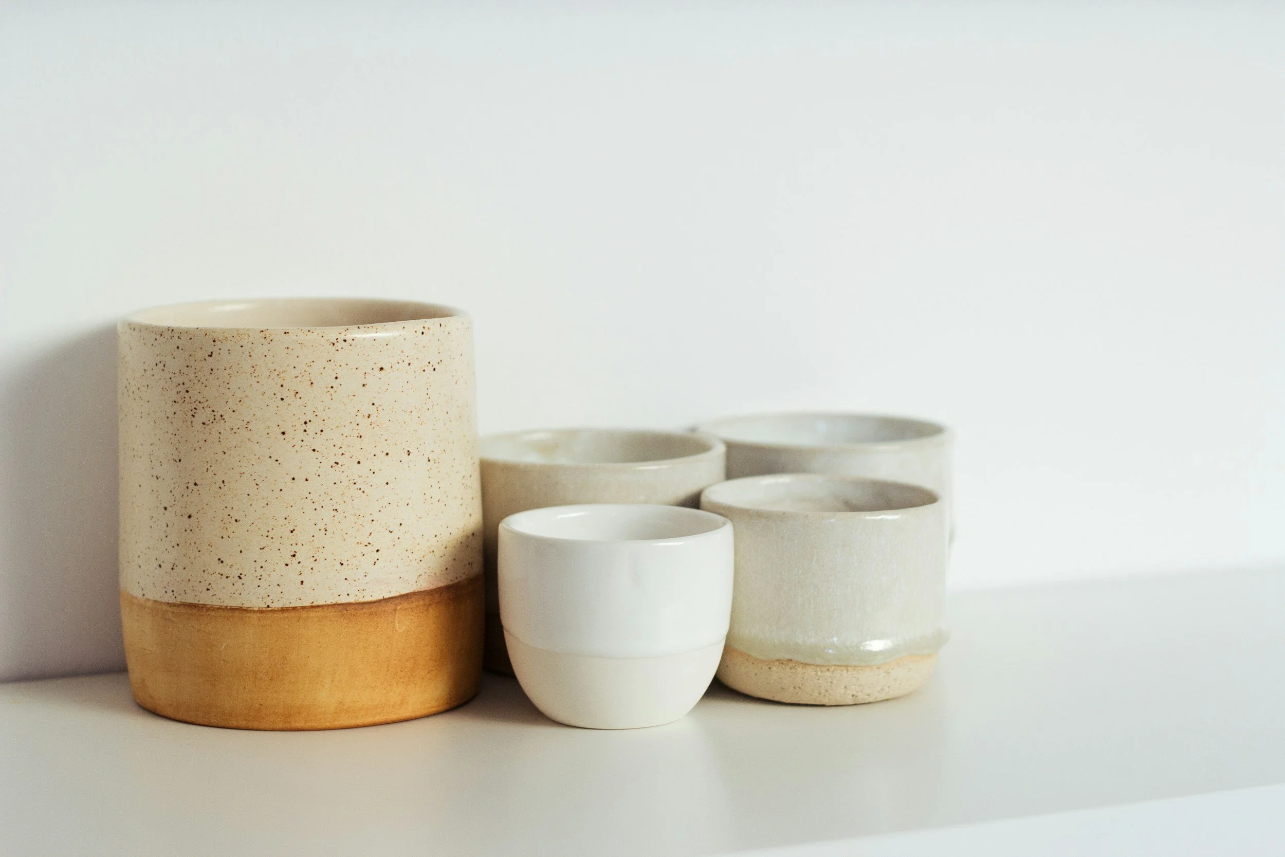 Set of five minimalistic ceramic cups, two larger speckled and textured cups in natural tones, and three smaller smooth white cups, arranged on a white surface against a plain white background.