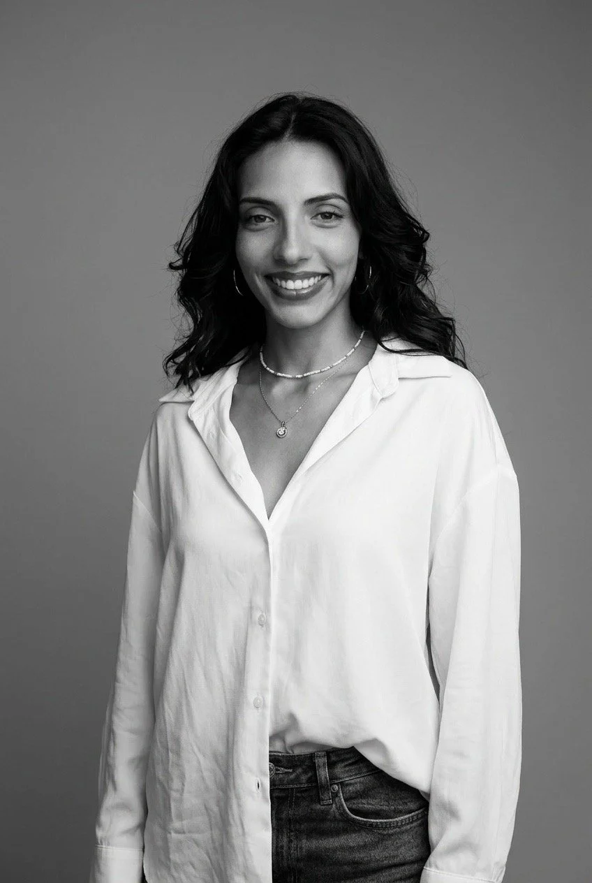 A black-and-white portrait of a woman with wavy dark hair, smiling, wearing a white button-up shirt, layered necklaces, and hoop earrings, standing against a plain background.