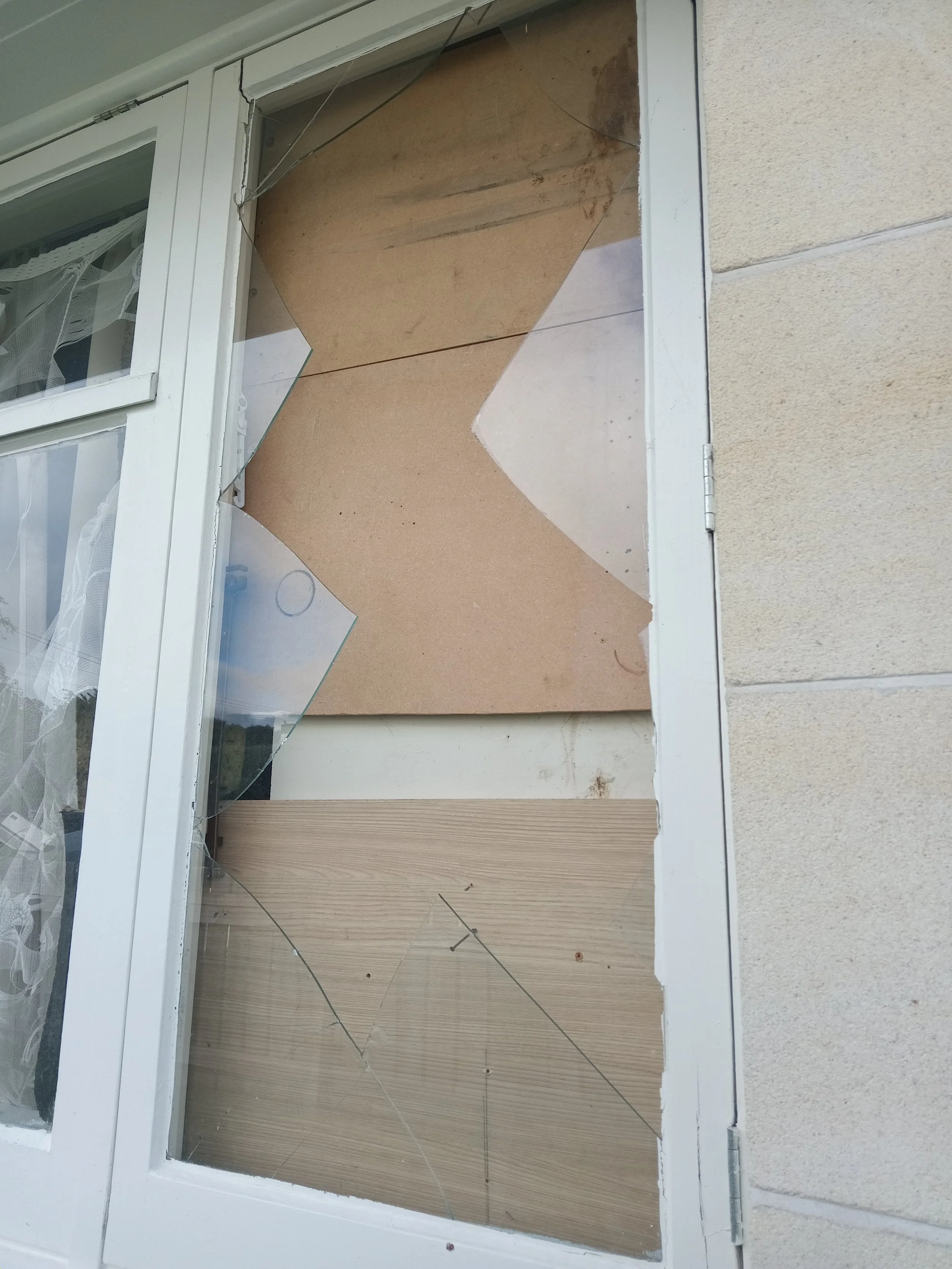 Broken glass pane of a sliding glass door with some panes missing, revealing the interior of a room with wood paneling, a window with white lace curtains, and a beige exterior wall.