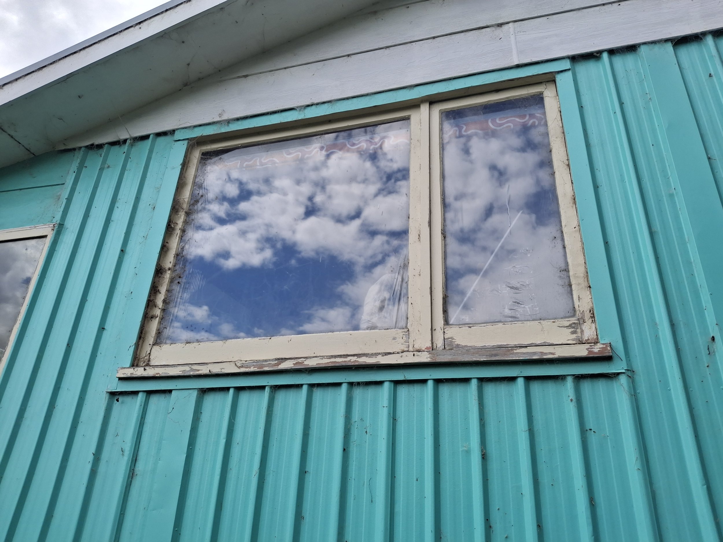 Before picture close-up of a window set, single glazed. The window has a white frame, and the panes reflect clouds and a blue sky. The window sill appears weathered and slightly dirty.