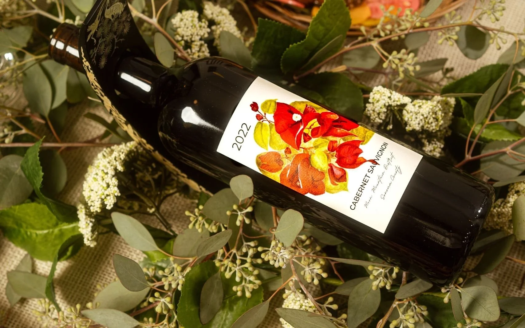 A bottle of Cabernet Sauvignon wine from 2022 lying on a bed of green leaves and white flowering plant.