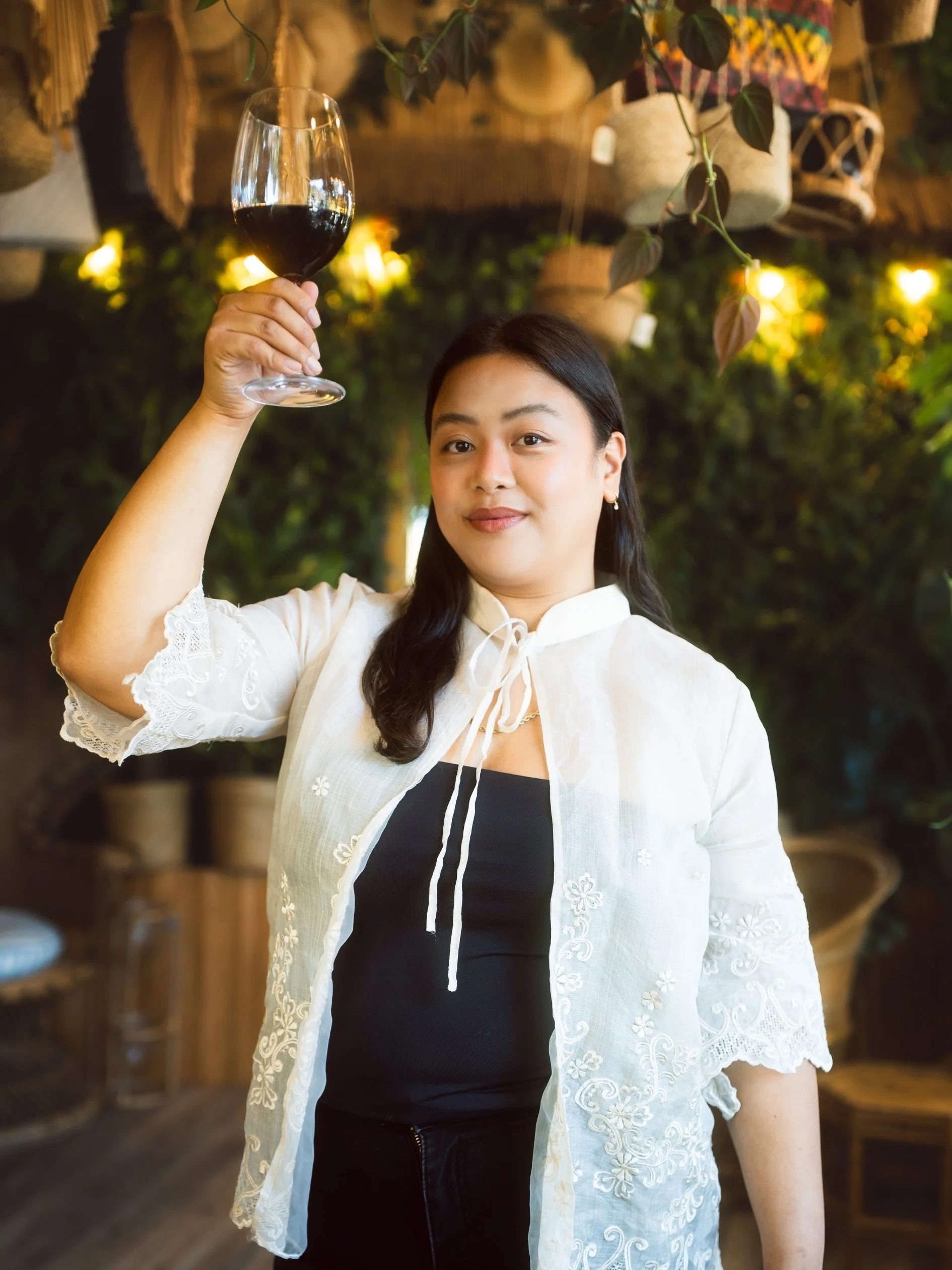 A woman in a white embroidered jacket holding a glass of red wine in an indoor setting with plants and warm lighting in the background.