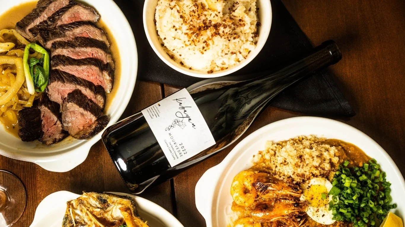 Assorted dishes of beef, shrimp, rice, and breaded shrimp, a bottle of red wine on a wooden table.