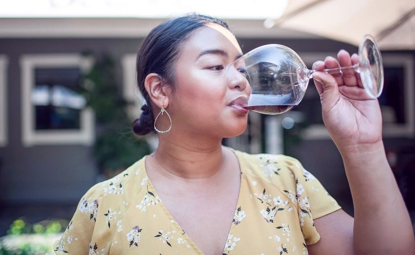Woman with dark hair in a bun, wearing earrings and a yellow floral dress, drinking red wine from a glass outdoors.