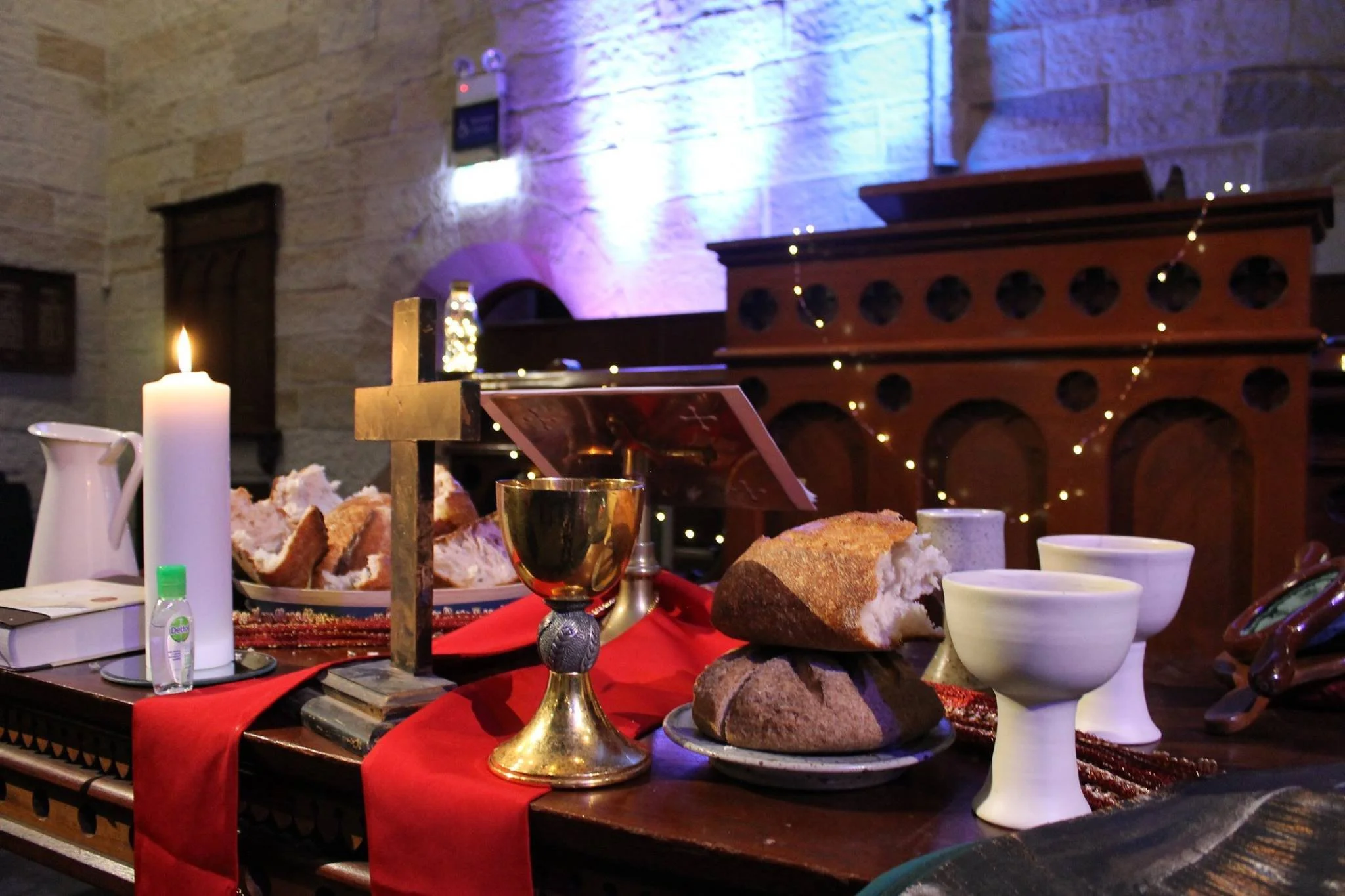 A religious altar decorated with a lit white candle, bread, wine, and cups, set on a red cloth with a small cross in the background, against a stone wall with purple and blue lighting.