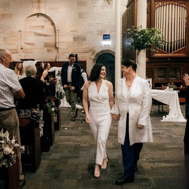 Two women holding hands and smiling at each other inside a church during a wedding ceremony, with guests seated on either side.