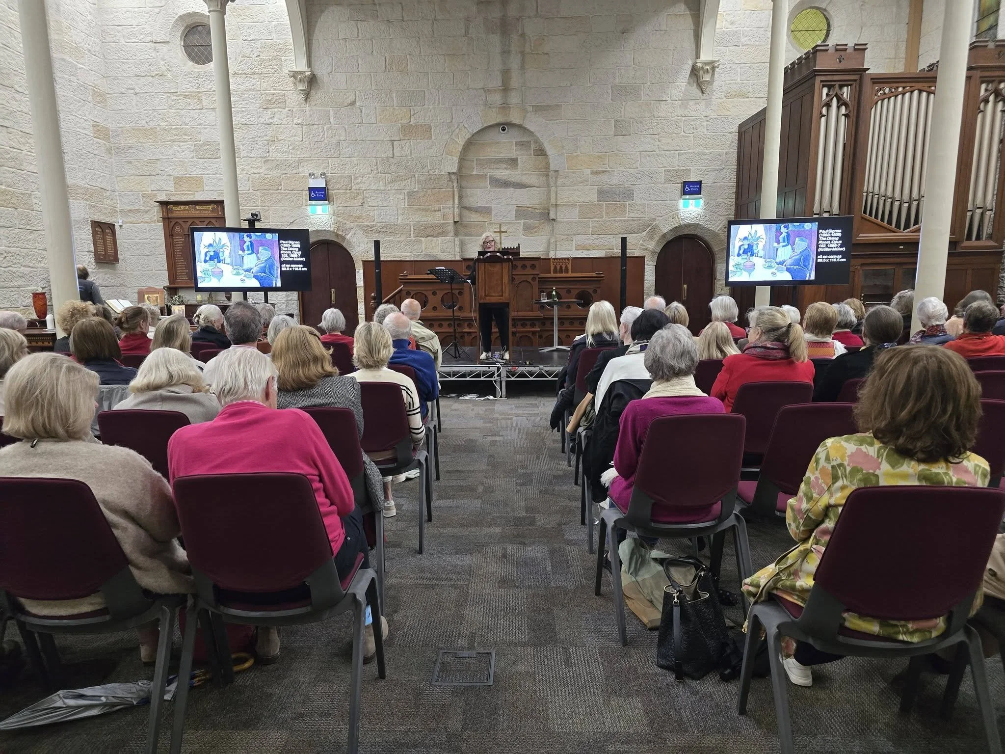 Inside a church or chapel with stone walls, an aisle, and wooden elements. A speaker stands at a podium in front of an audience of older adults, some wearing glasses and colorful clothing. Two display screens show an artwork of people at a table in a room with a pineapple plant in the background.