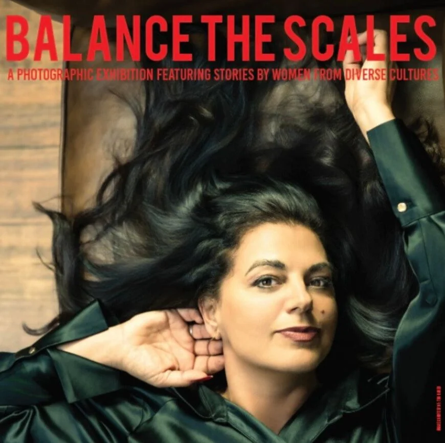 Poster for a photography exhibition titled 'Balance the Scales' featuring stories by women from diverse cultures, showing a woman with dark hair lying down and looking at the camera.