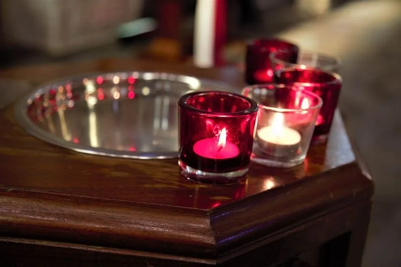 Three lit candles in red and clear glass holders on a wooden table with a reflective silver tray nearby.