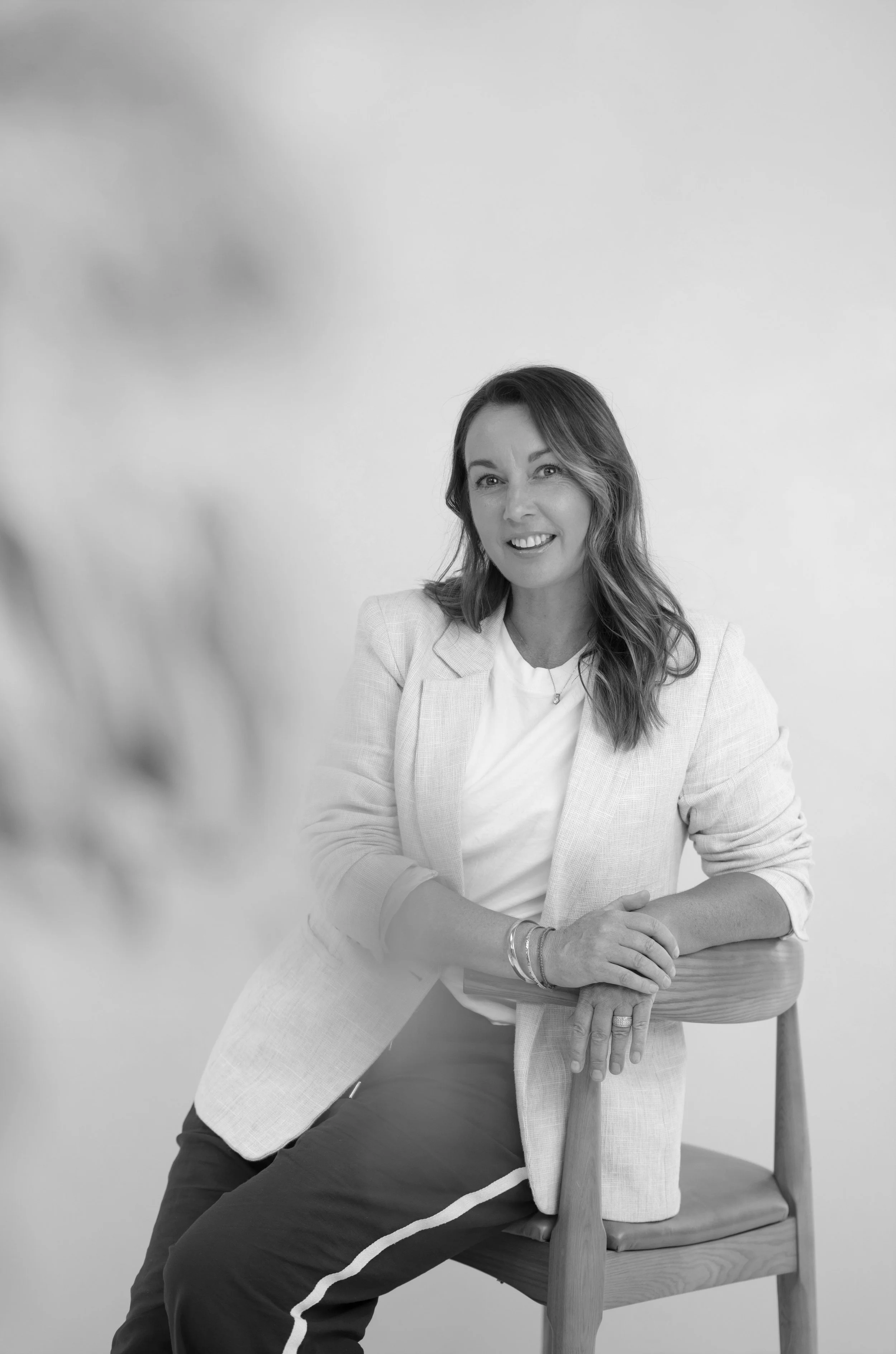 Black and white photo of a woman with long wavy hair, wearing a light blazer and a white top, sitting on a wooden chair, smiling at the camera, with her left arm resting on the table and her right arm on her lap.