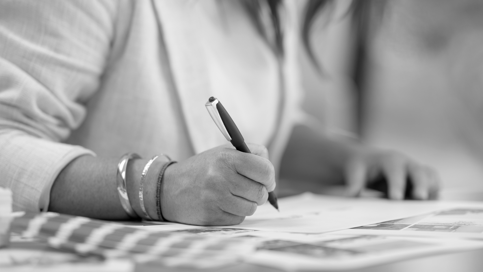 A person writing on a paper with a pen, wearing bracelets and a long sleeve shirt, photographed in black and white.