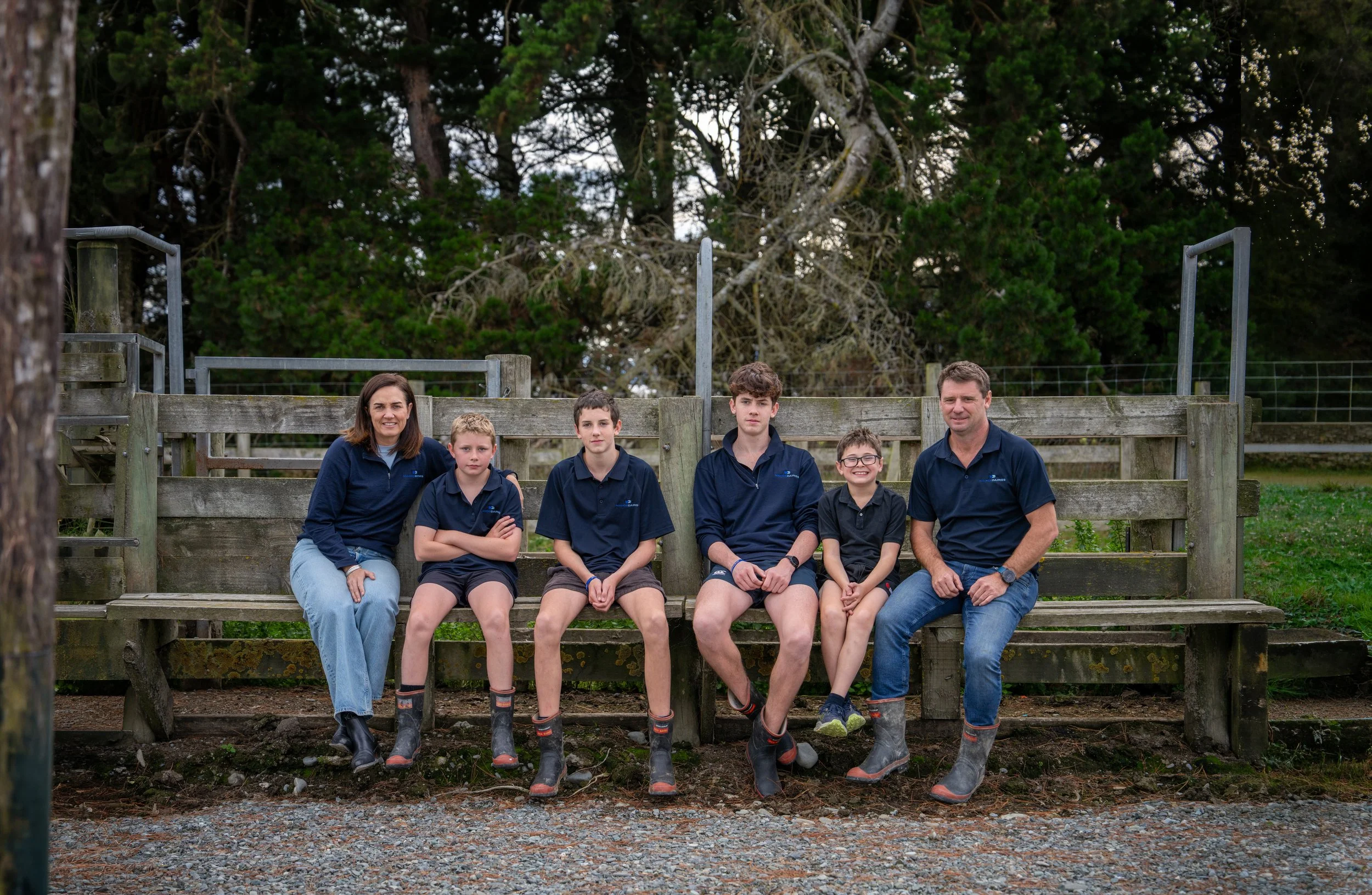 Family of six sitting on a wooden bench outdoors, with a backdrop of trees and green foliage. They are dressed in casual clothing, with four wearing matching navy polo shirts, and all are smiling at the camera.