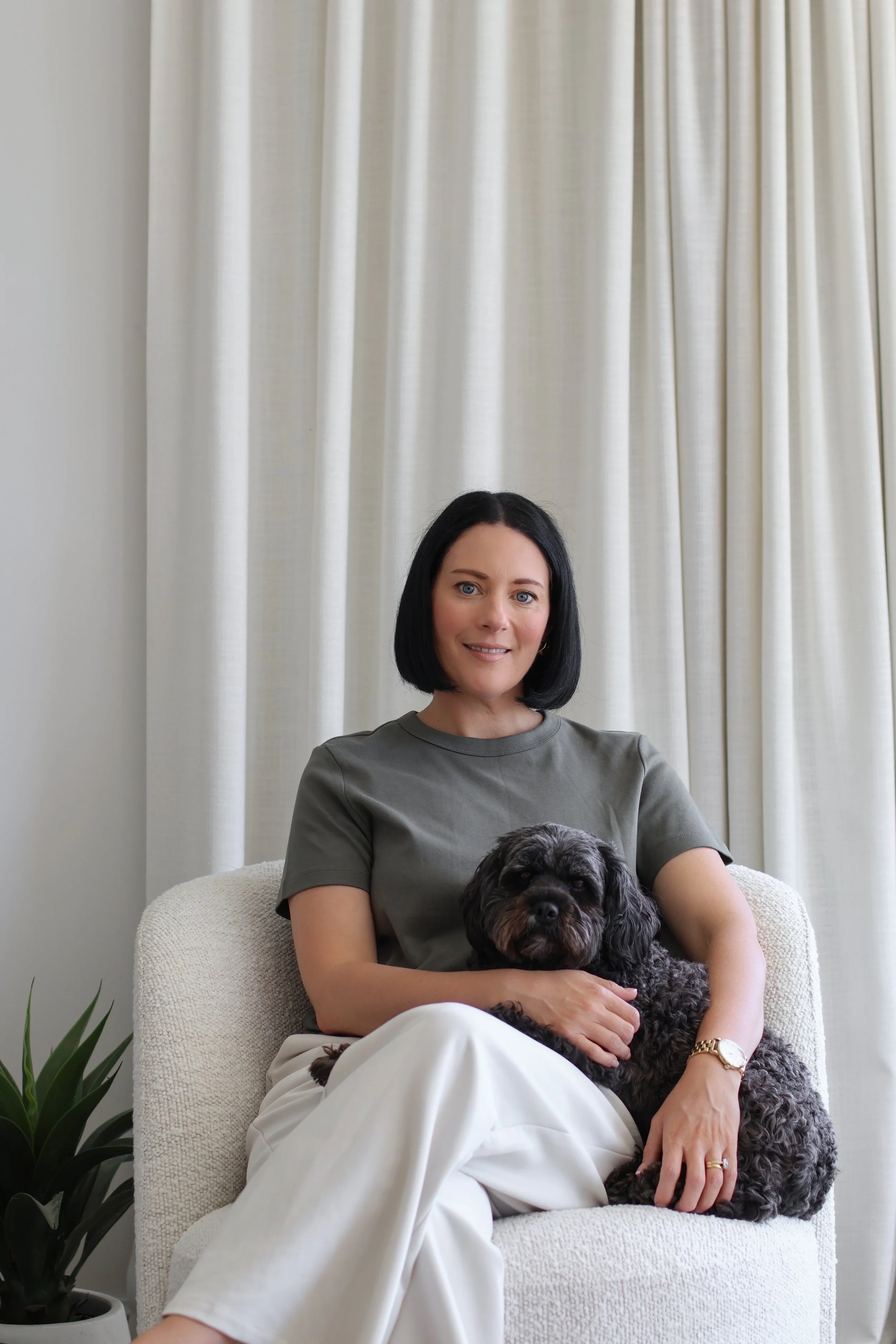 A woman with black hair, wearing a gray shirt, sitting on a cream-colored armchair, holding a black and brown dog on her lap, with a white curtain in the background.
