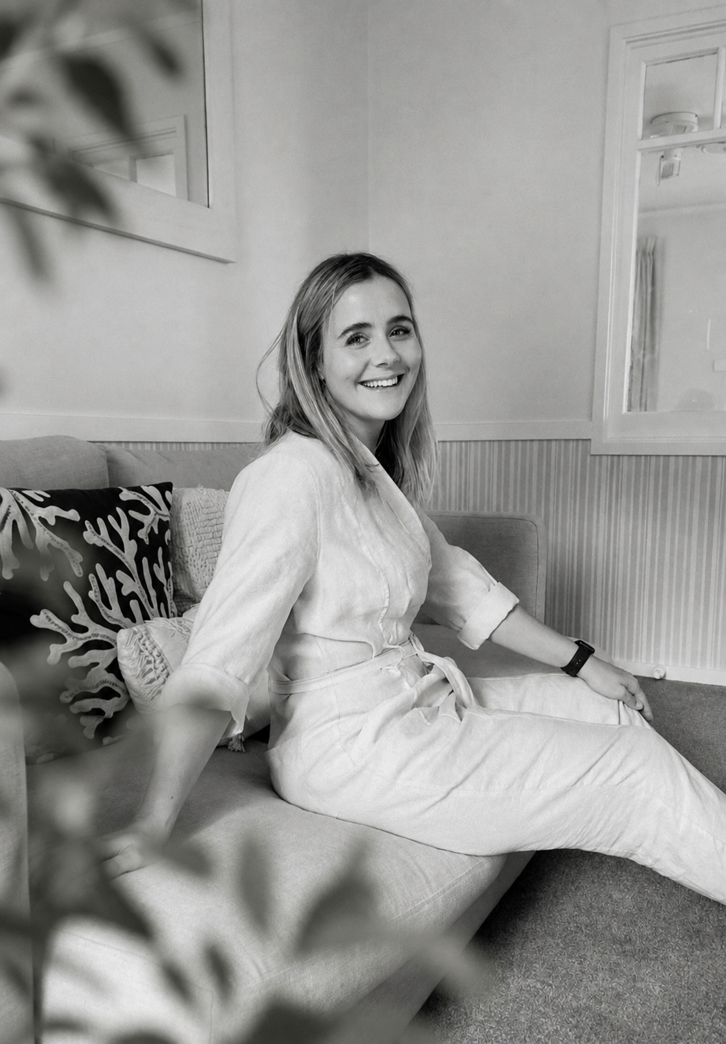 A smiling woman with light hair wearing light-colored pajamas sits on a sofa, looking at the camera in a cozy room with cushions and framed artwork on the wall.