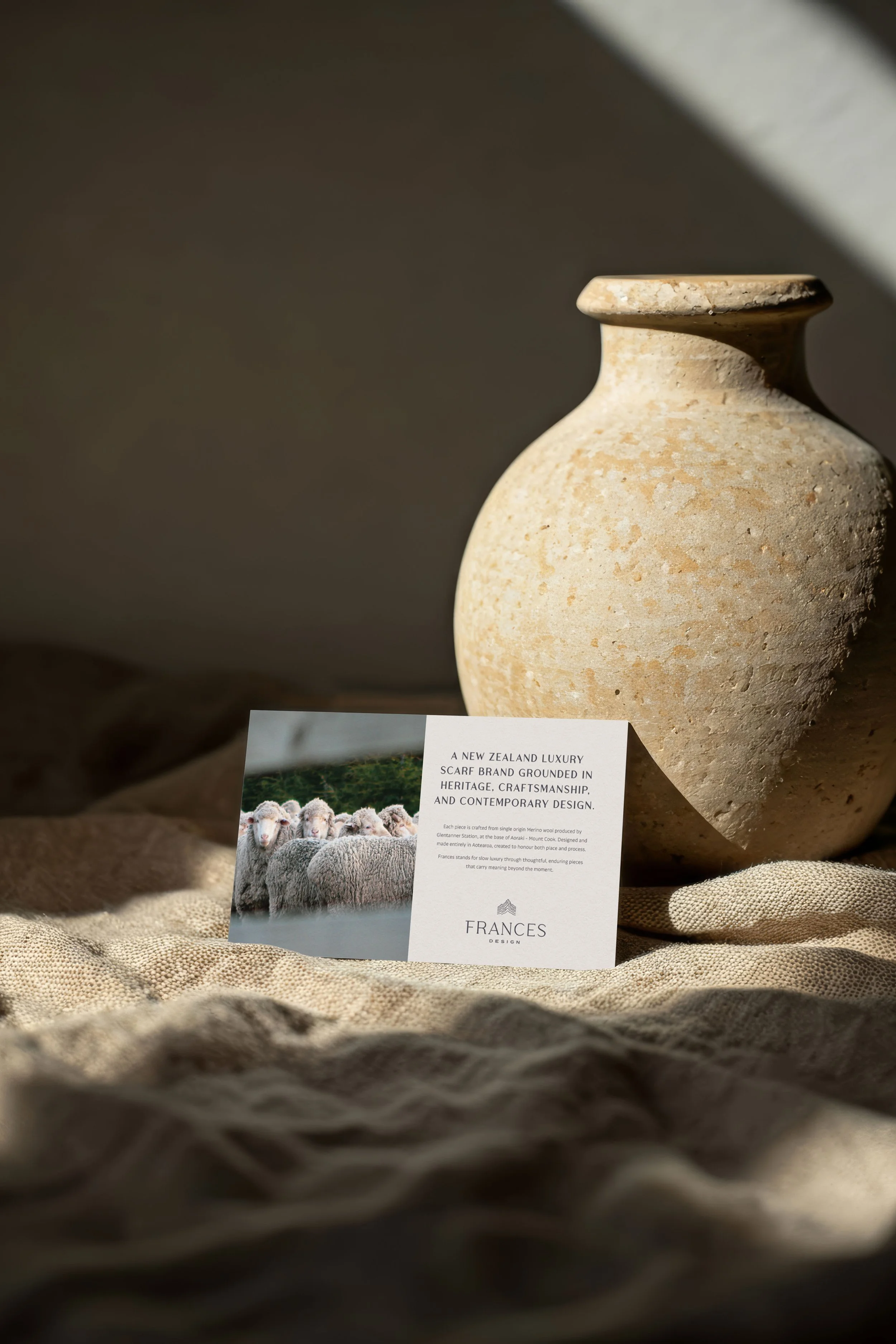 A beige ceramic vase with a rounded body and short neck placed on a textured surface with a piece of paper in front of it. The paper has a photograph of sheep and text about a New Zealand luxury scarf brand called Franges Design.
