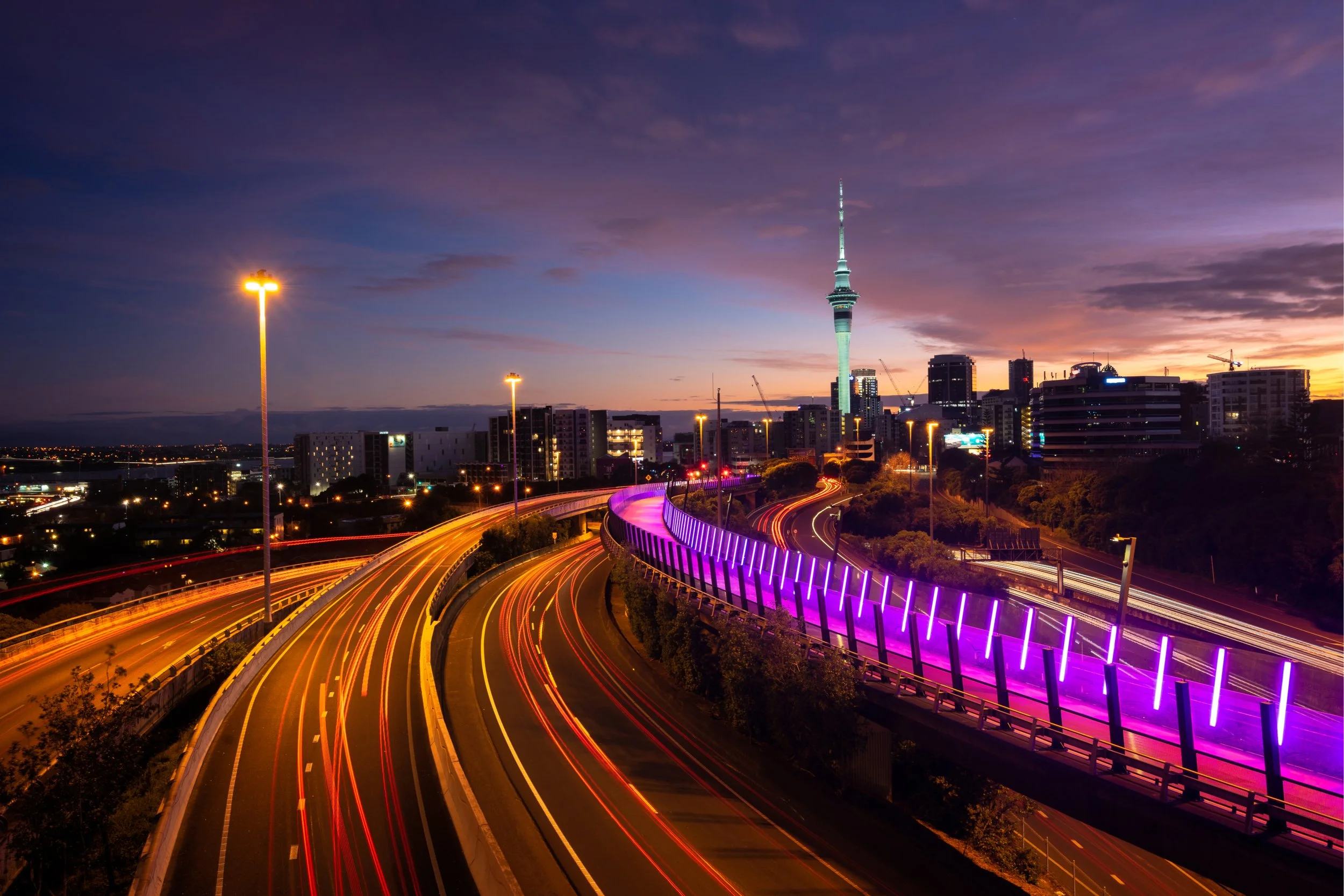 Auckland skyline at dusk featuring the illuminated Sky Tower, city buildings, and light trails on curving highways.
