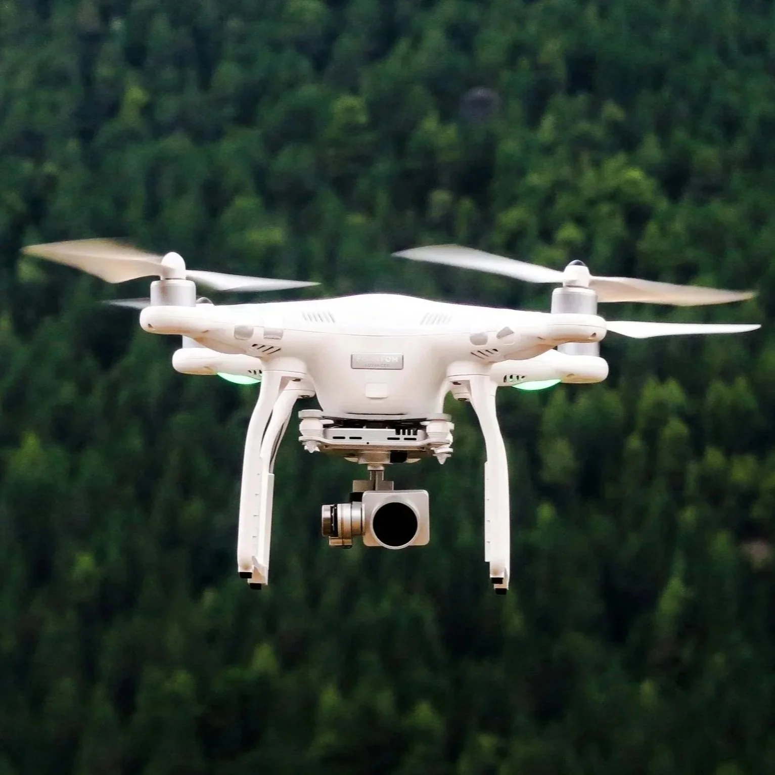 White quadcopter drone with mounted camera flying in front of a blurred forest