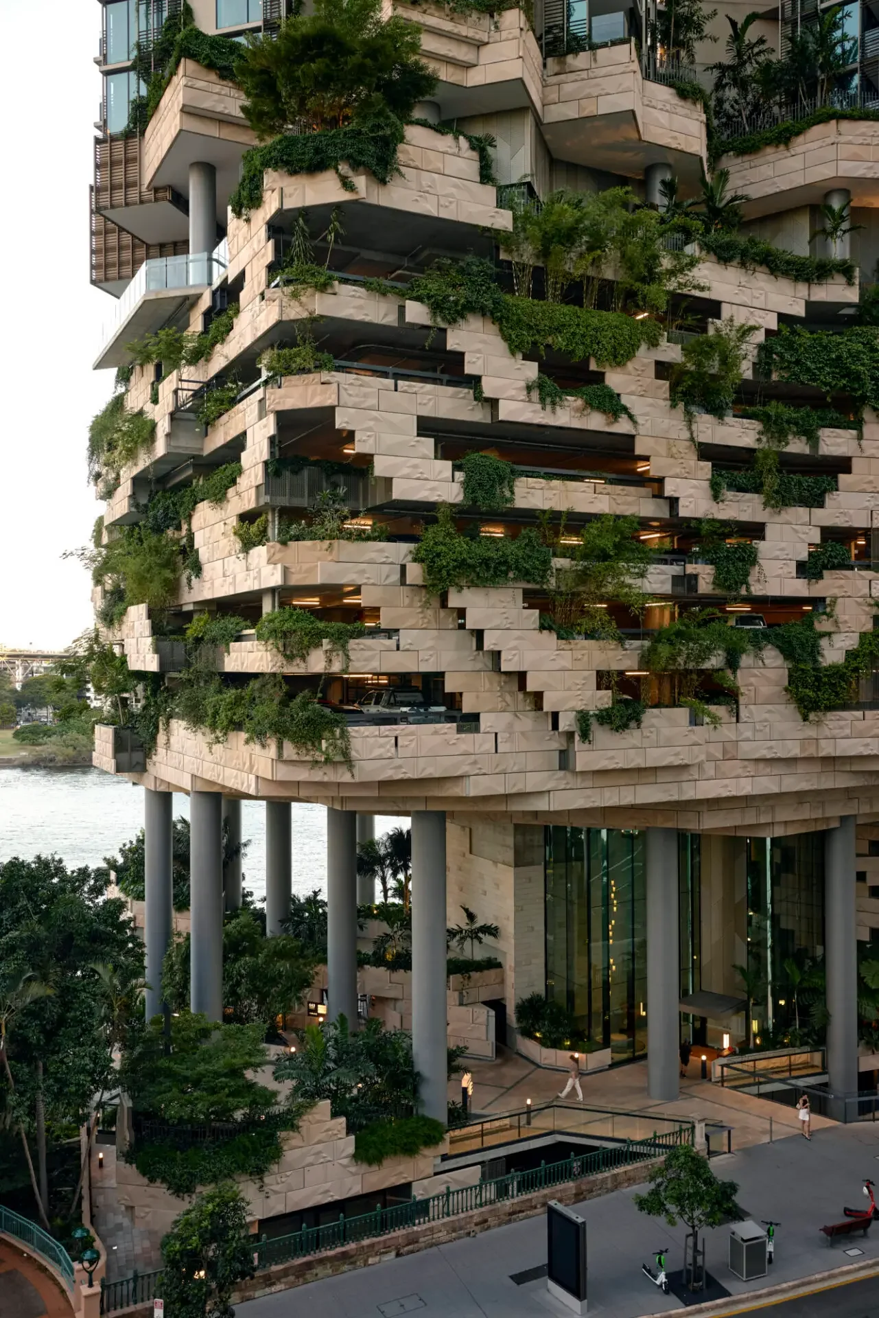 Modern waterfront building with textured stone terraces, lush greenery, balconies, and tall columns