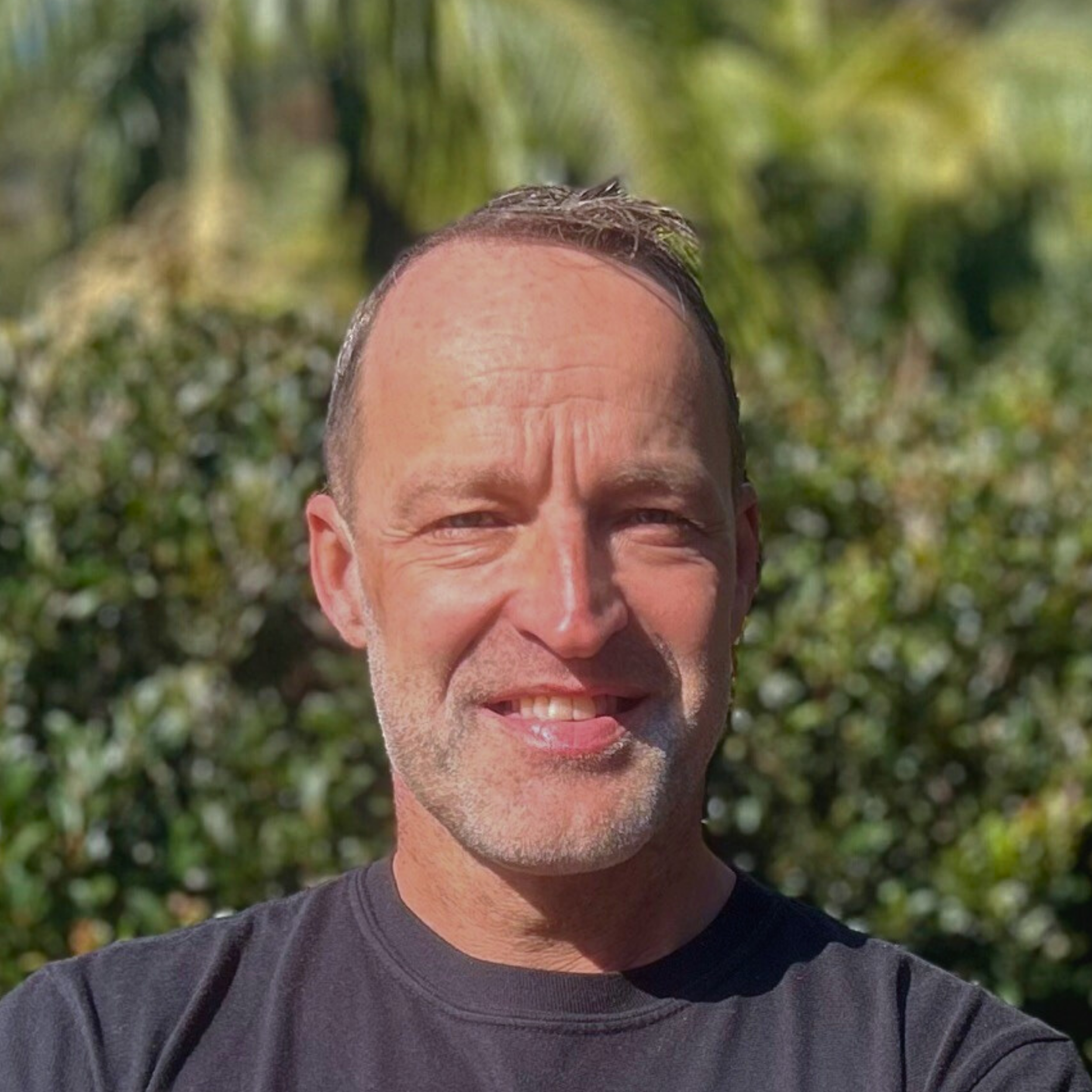 Smiling man in a dark T-shirt outdoors with leafy greenery in the background