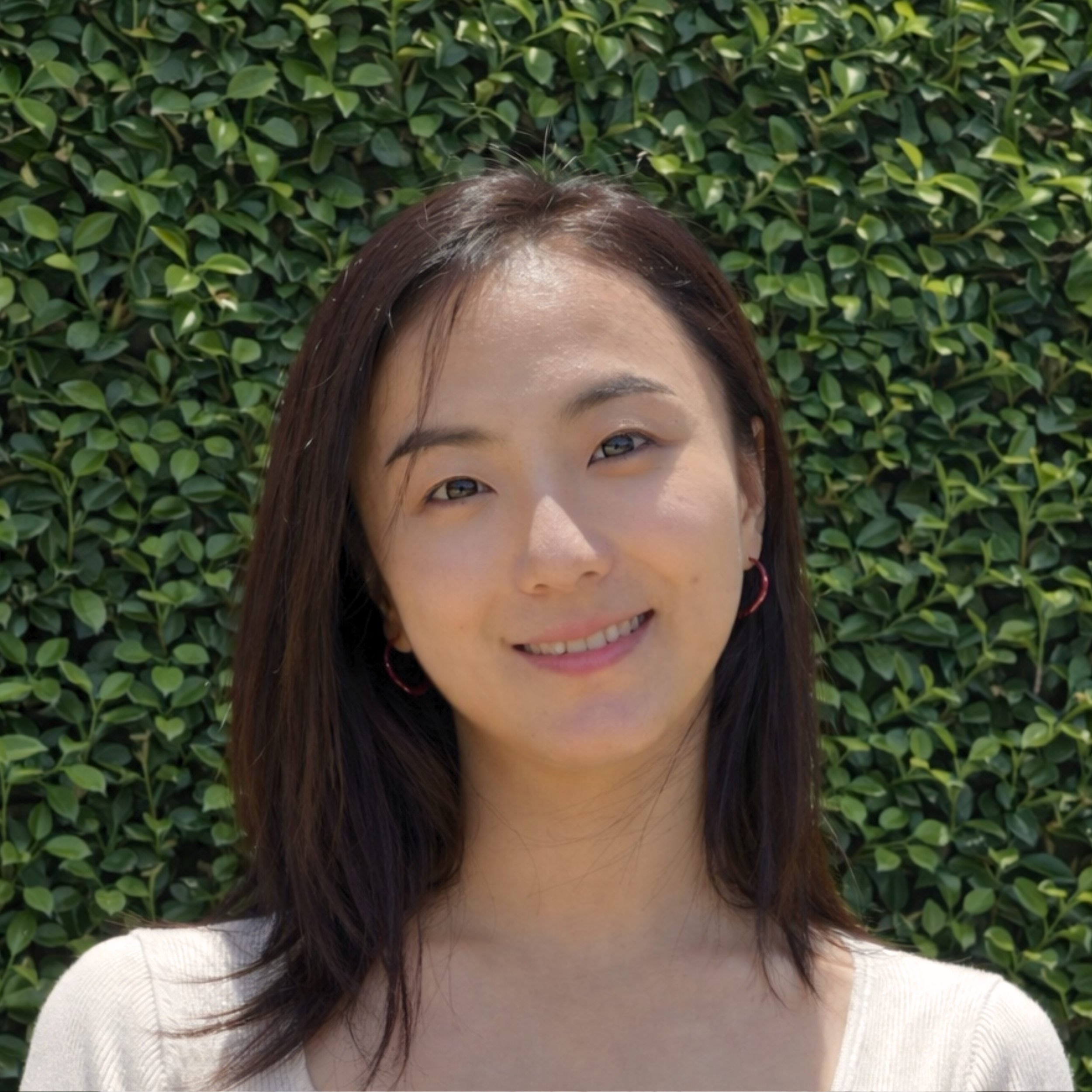 Smiling woman with long brown hair and hoop earrings in front of a green hedge